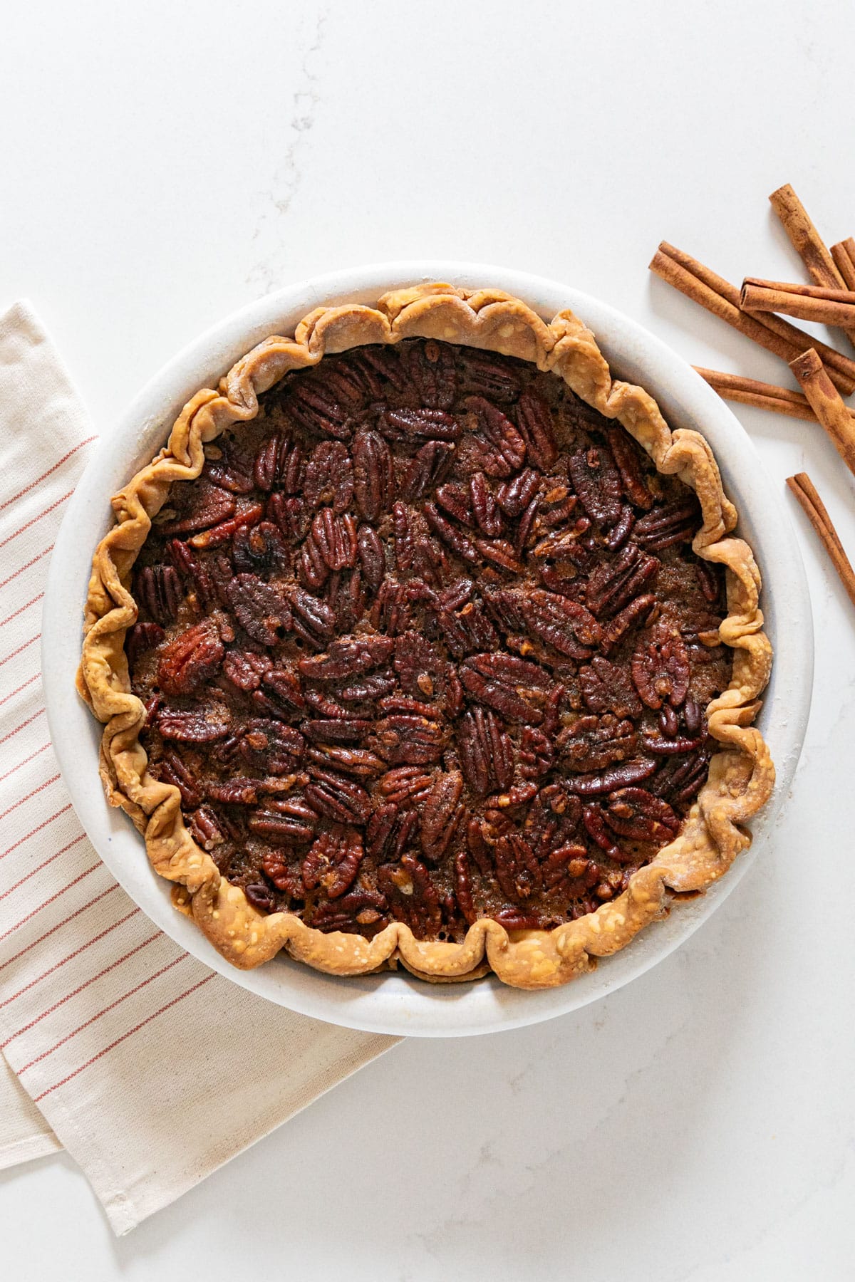 An overhead image of a pecan pie with a striped napkin and cinnamon sticks next to it.