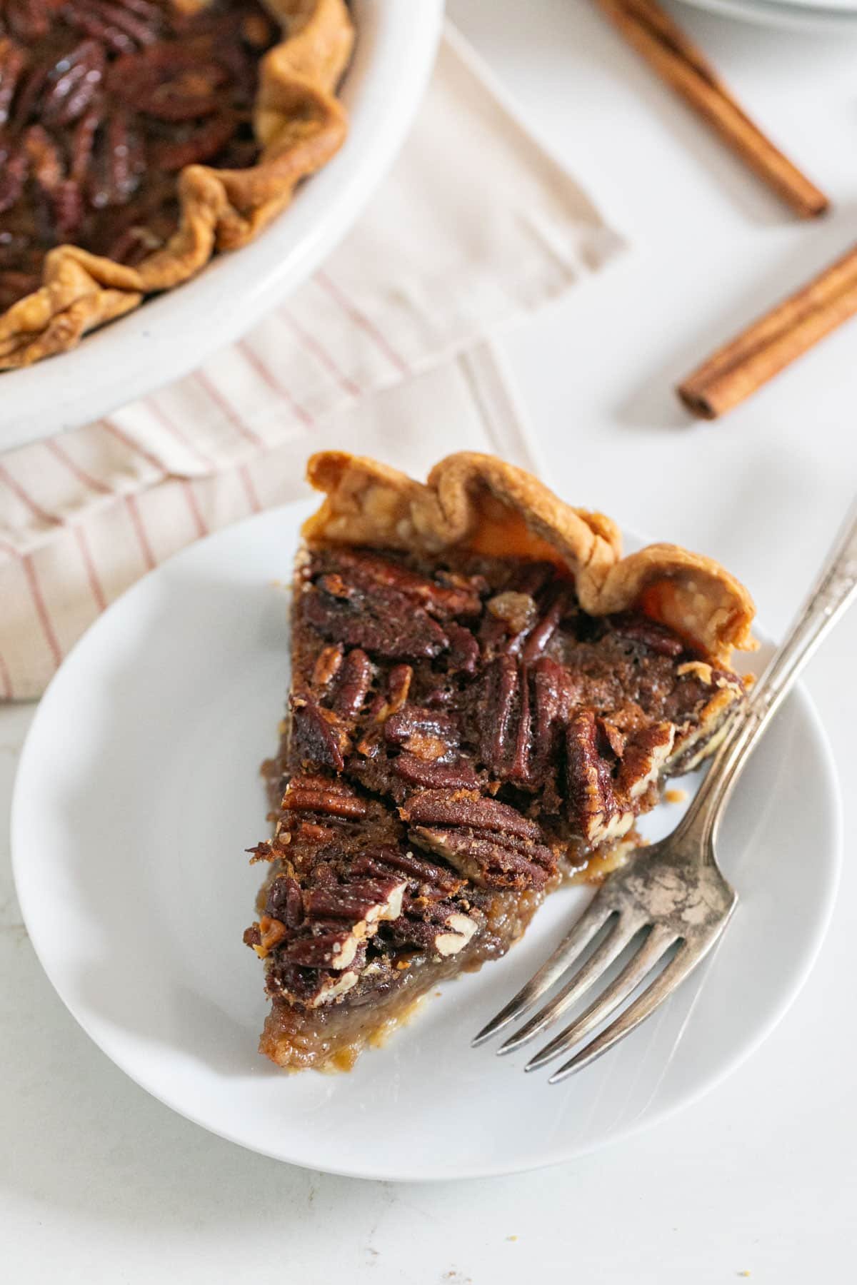A slice of pecan pie on a white plate with a fork on it and the rest of the pie behind it.