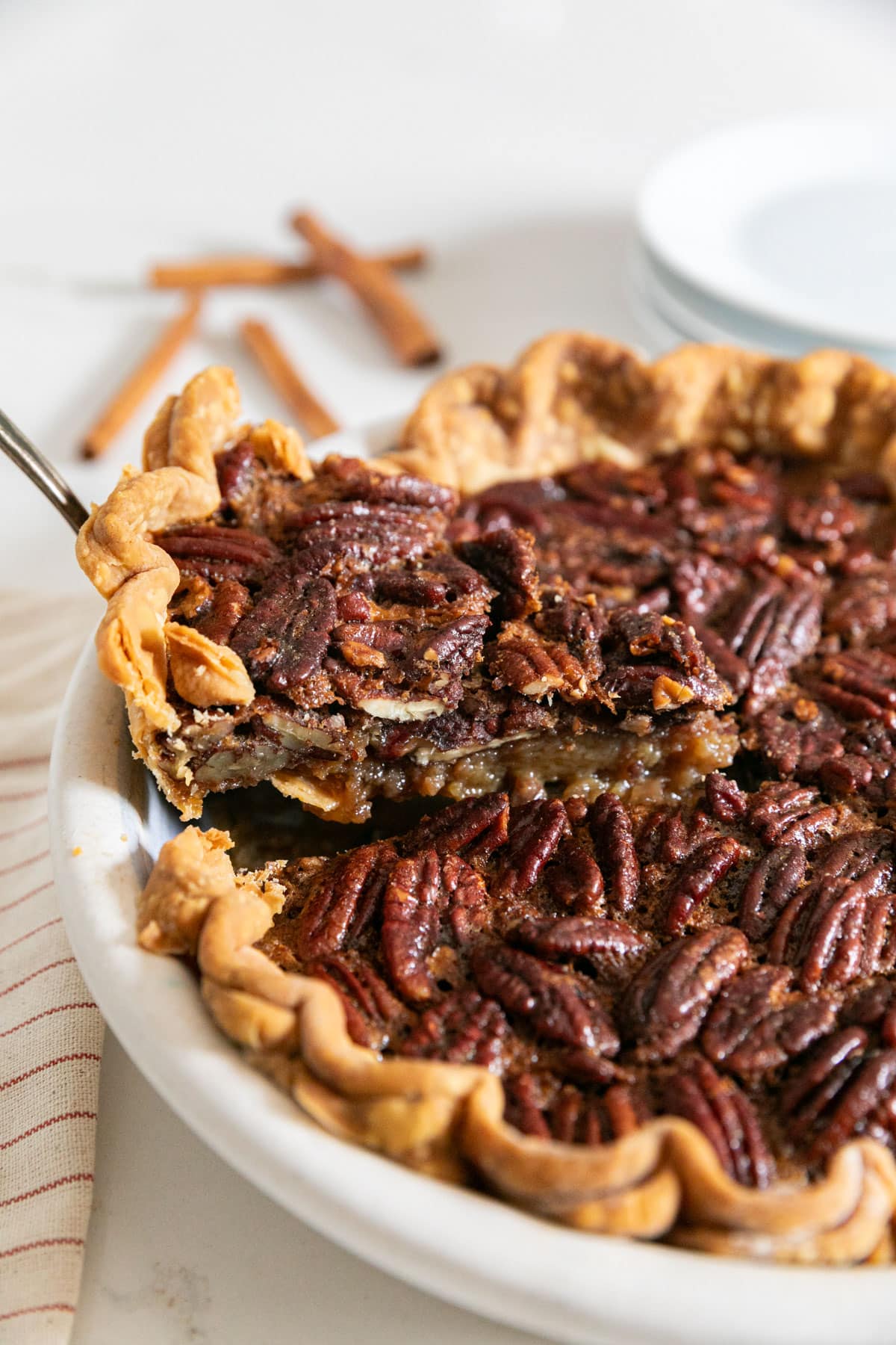 A slice of pecan pie being pulled from the pie with plates and cinnamon sticks in the background.