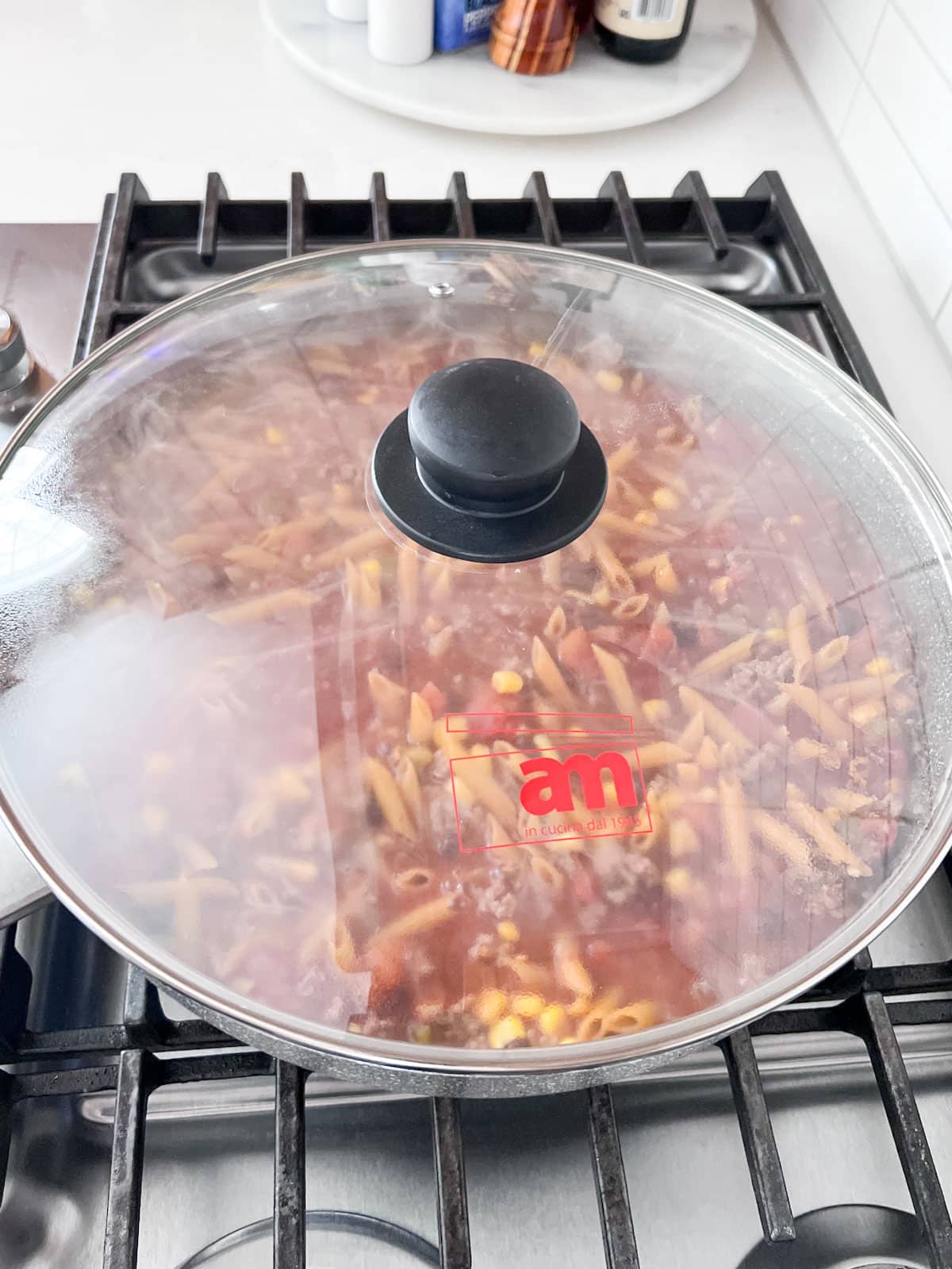 The pasta in a skillet on a stove cooking with a clear cover on it.