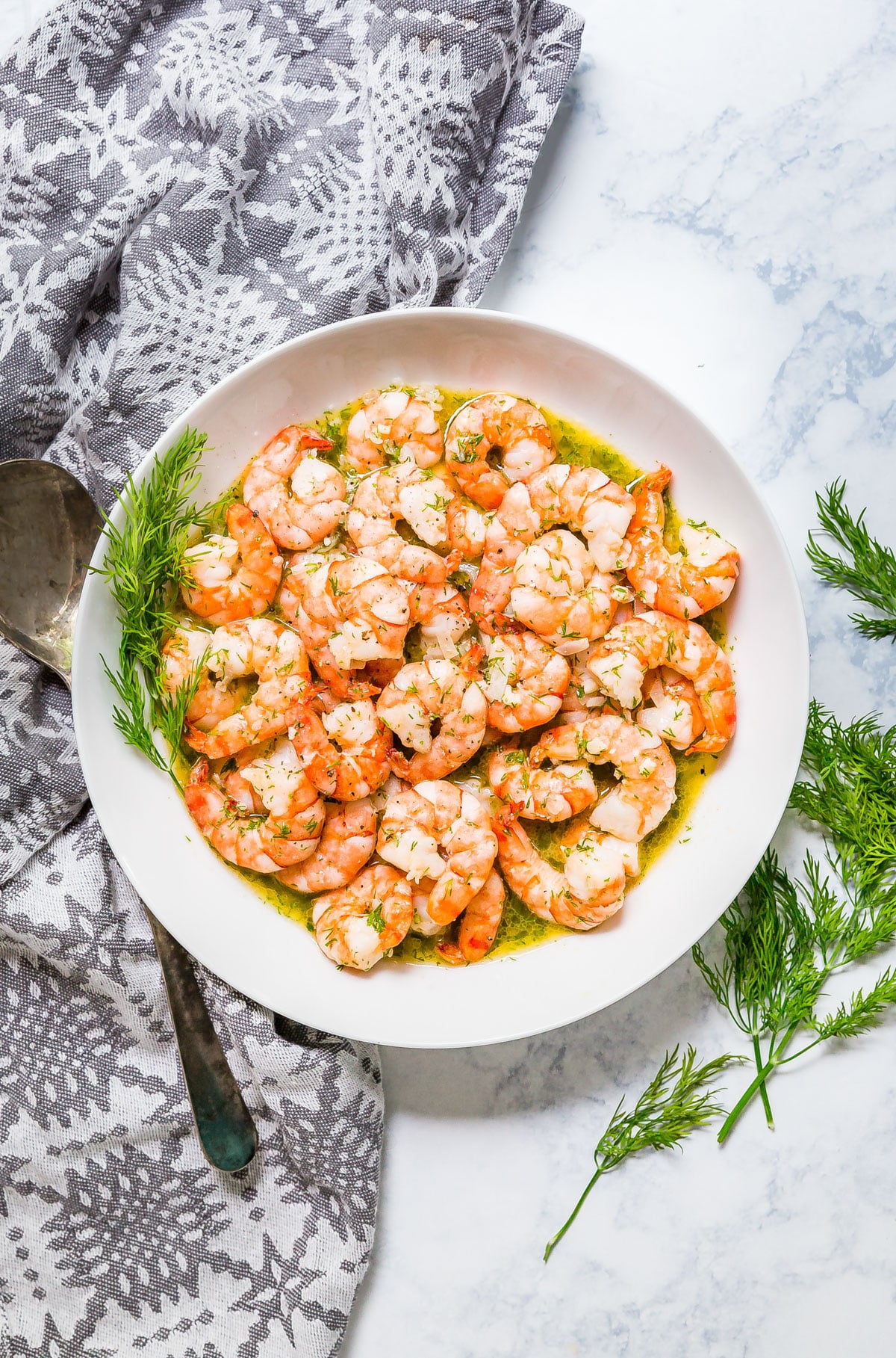 An overhead image of a white bowl of sautéed shrimp with fresh dill and a grey napkin around it.