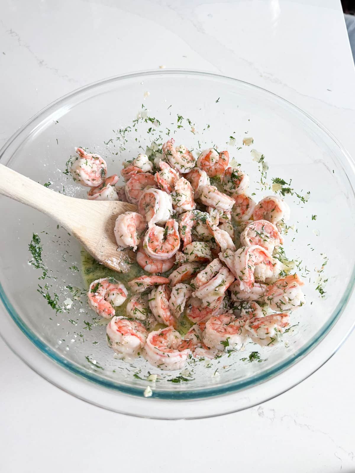 Shrimp in a glass bowl with dill and lemon juice on it being stirred with a wooden spoon.