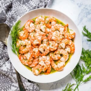 An overhead image of a white bowl of sautéed shrimp with fresh dill and a grey napkin around it.