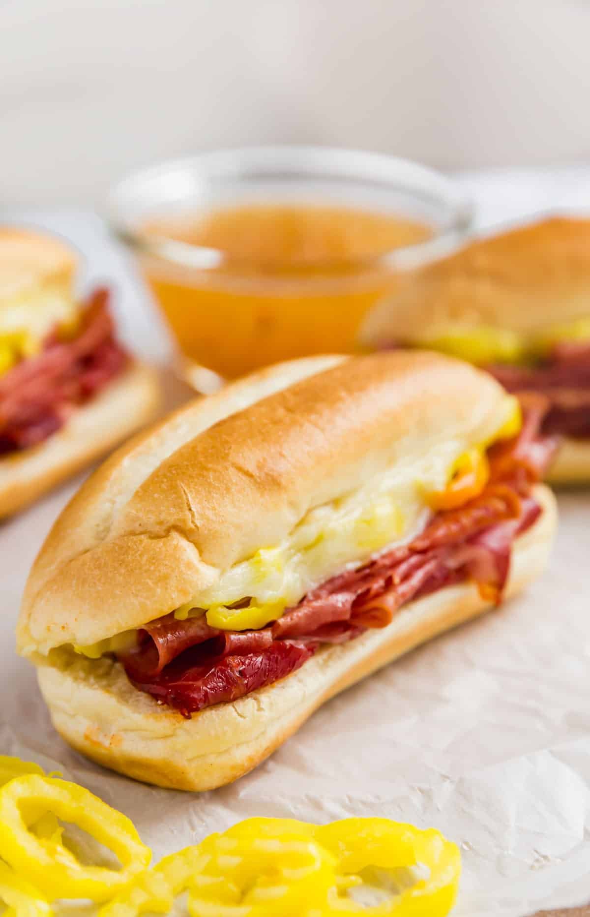 An image of an Italian sub sandwich with some banana peppers in front of it with other sandwiches and a bowl of Italian dressing in the background.