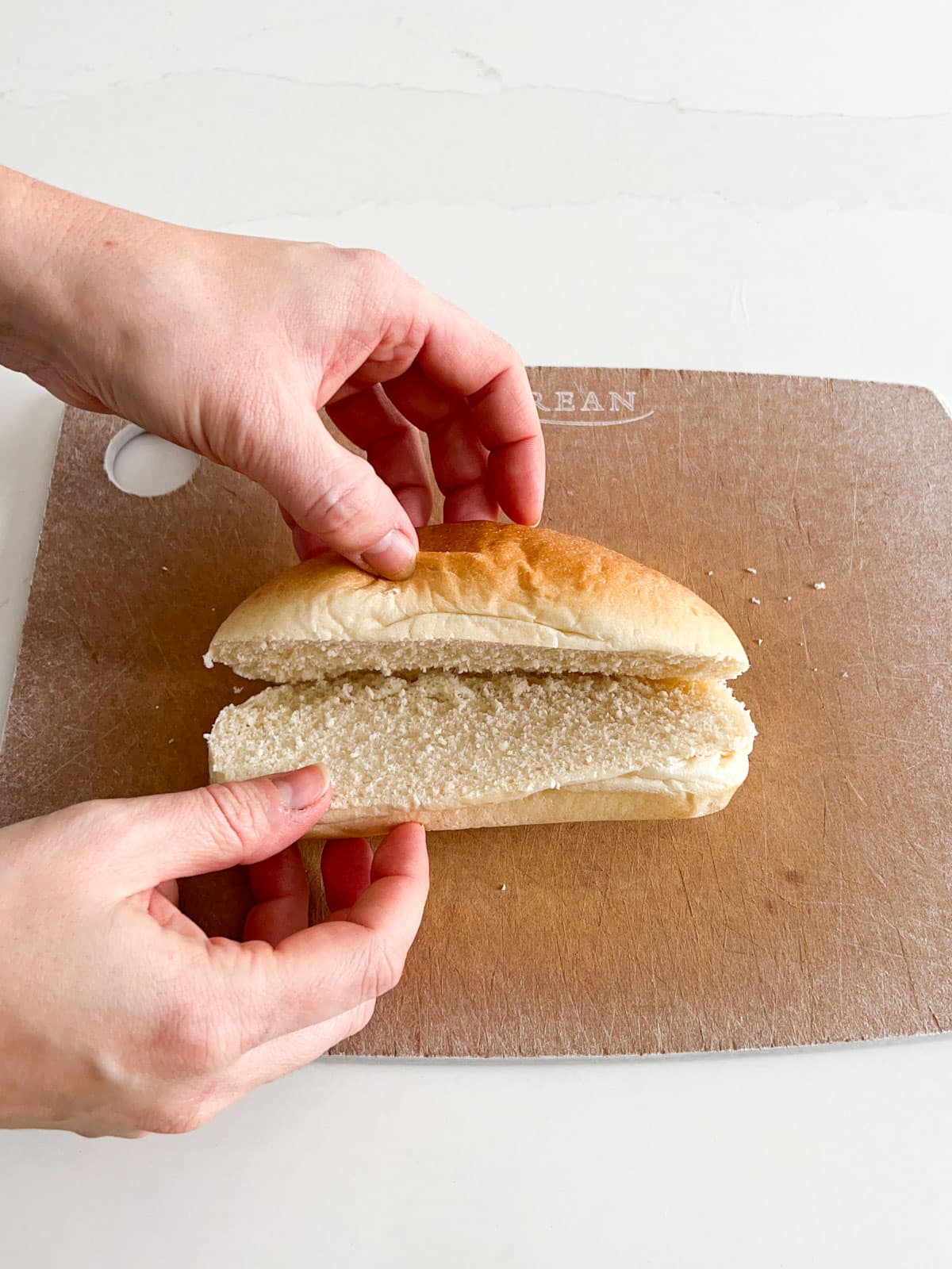 Hands holding a sub roll open on a cutting board  showing that it's still connected in the back.