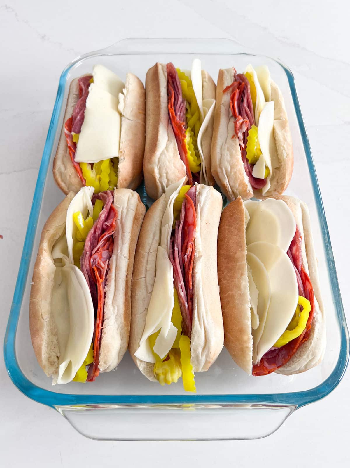 Grinder sandwiches filled with meats, cheese and banana peppers in a glass pan