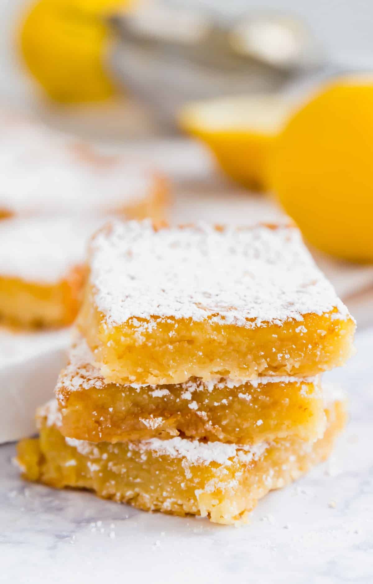 A stack of 3 lemon squares with lemons behind them.
