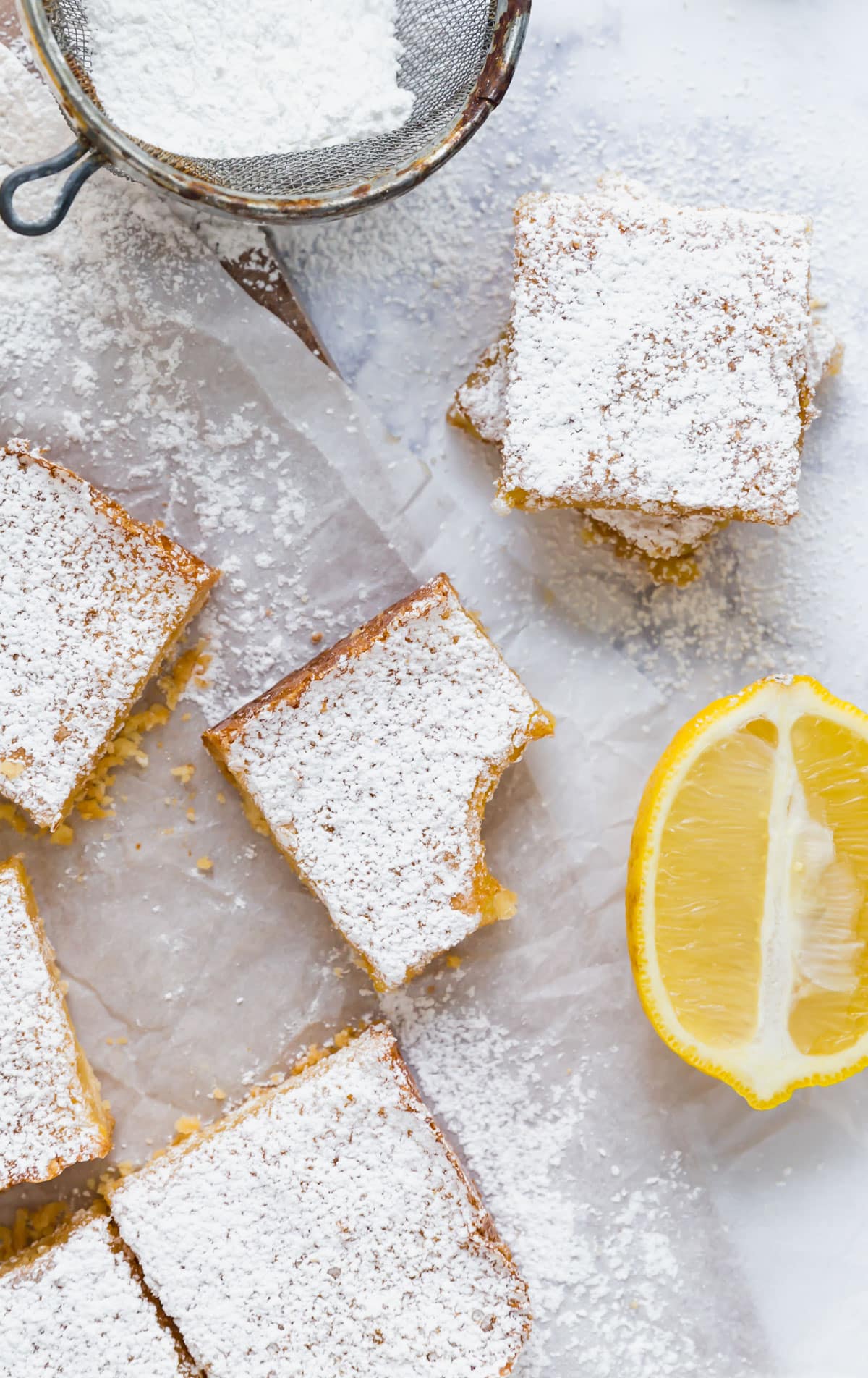 An overhead image of this lemon bar recipe cut into squares and scattered with one that has a bite out of it and a half lemon.