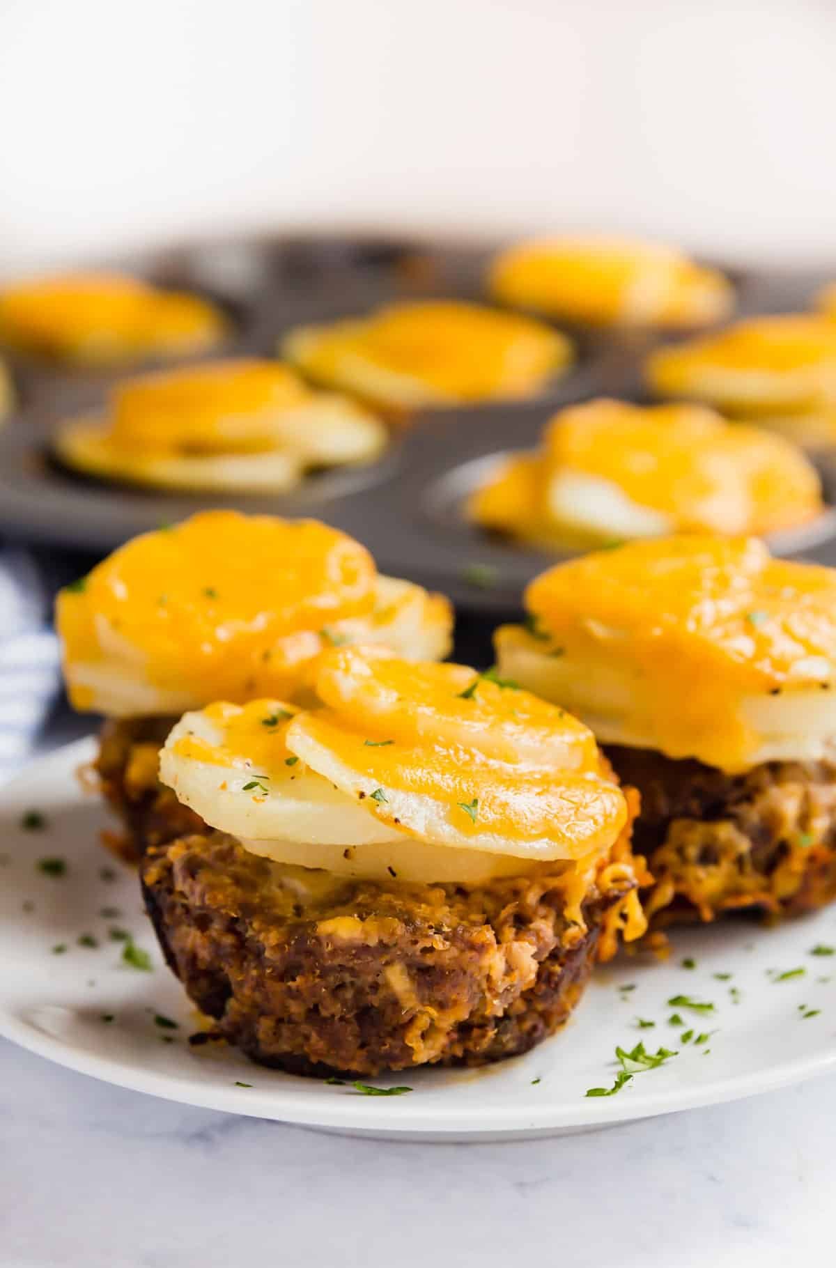 3 Mini Meatloaf Muffins topped with cheesy potatoes on a white plate with the pan behind it.