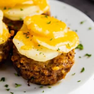 A close up of a meatloaf muffin topped with cheesy potatoes on a white plate.