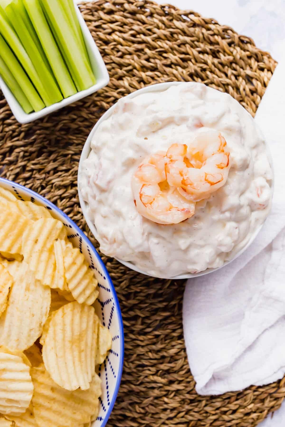 An overhead image of shrimp dip with a whole shrimp on top next to a bowl of chips and a bowl of celery sticks.