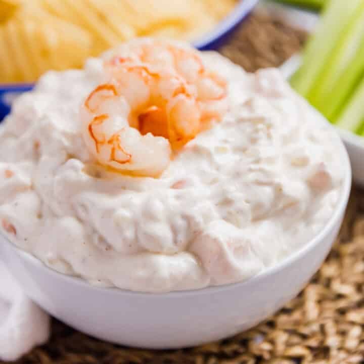 A close up image of a white bowl of shrimp dip with a whole shrimp on top and chips and celery in the background.