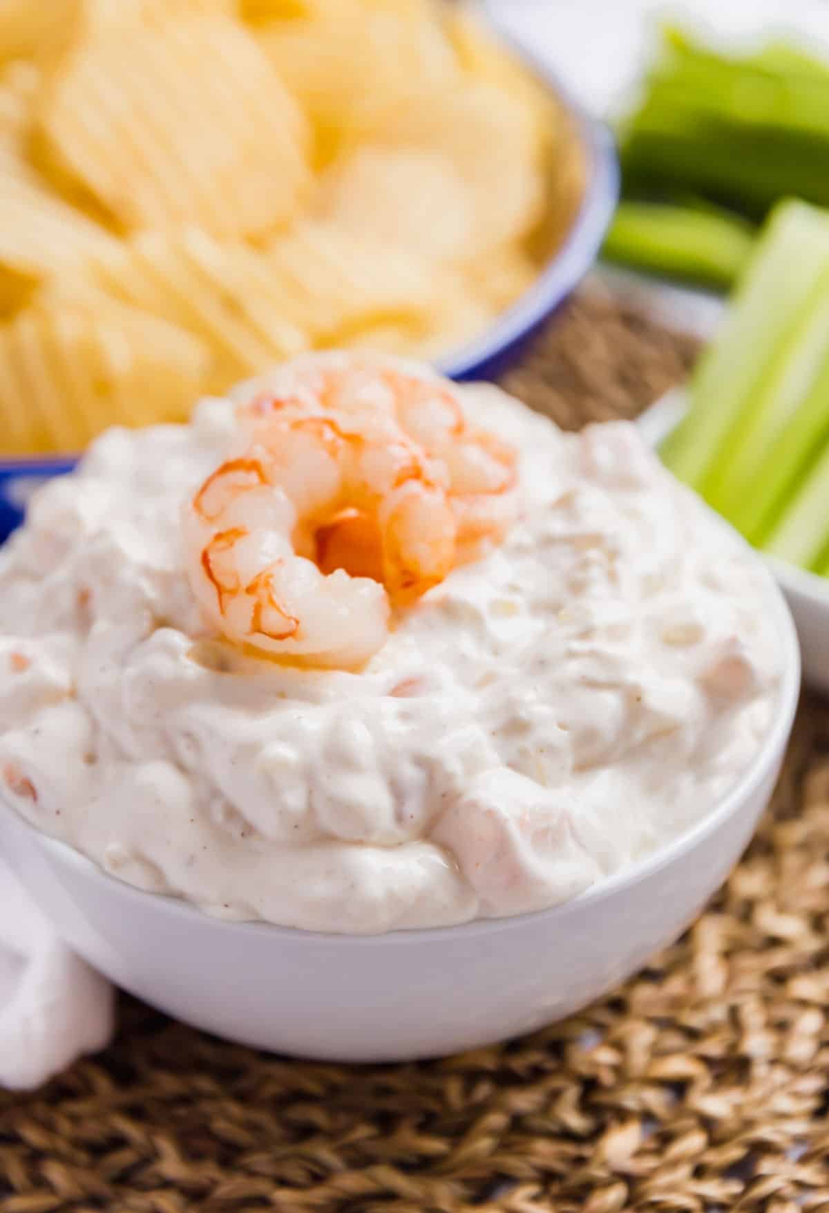 A close up image of a white bowl of shrimp dip with a whole shrimp on top and chips and celery in the background.