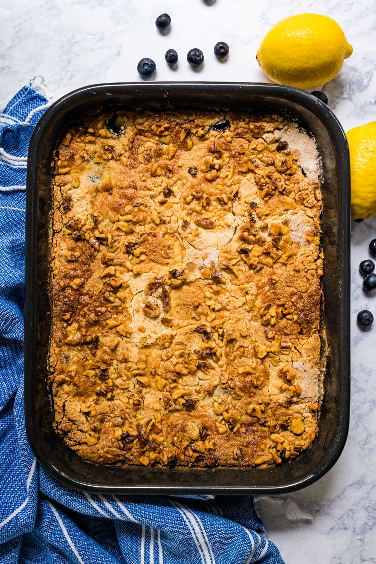 An overhead image featuring the top golden crust of the dump cake in a dark pan with a blue and white striped towel, blueberries and lemons around it.