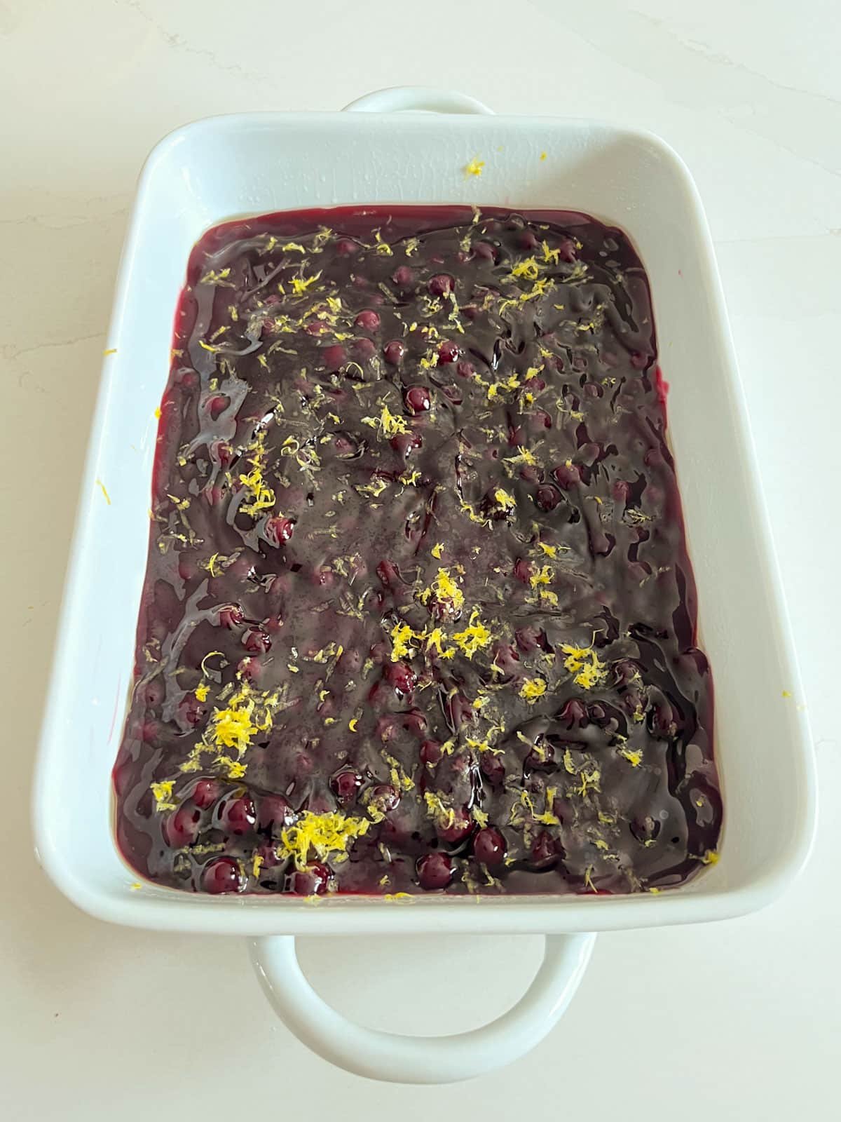 A white baking pan with blueberry pie filling spread in an even layer and topped with lemon zest.