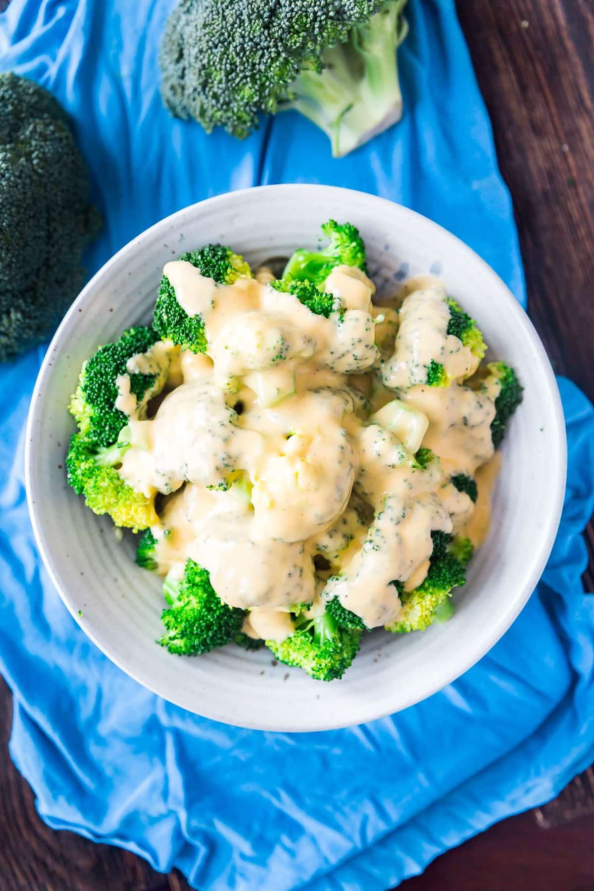 An overhead image of a bowl of steamed broccoli topped with homemade cheese sauce in a gray bowl on a blue napkin.