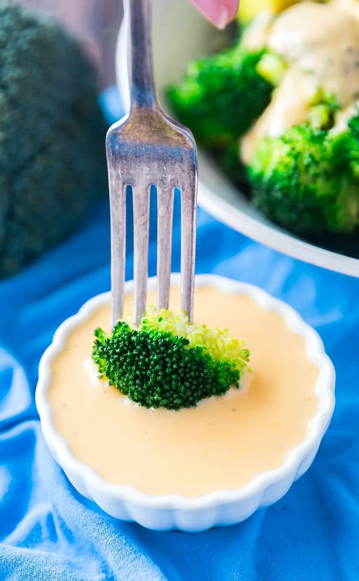 A fork dipping a piece of broccoli into a small white bowl of cheddar cheese sauce.