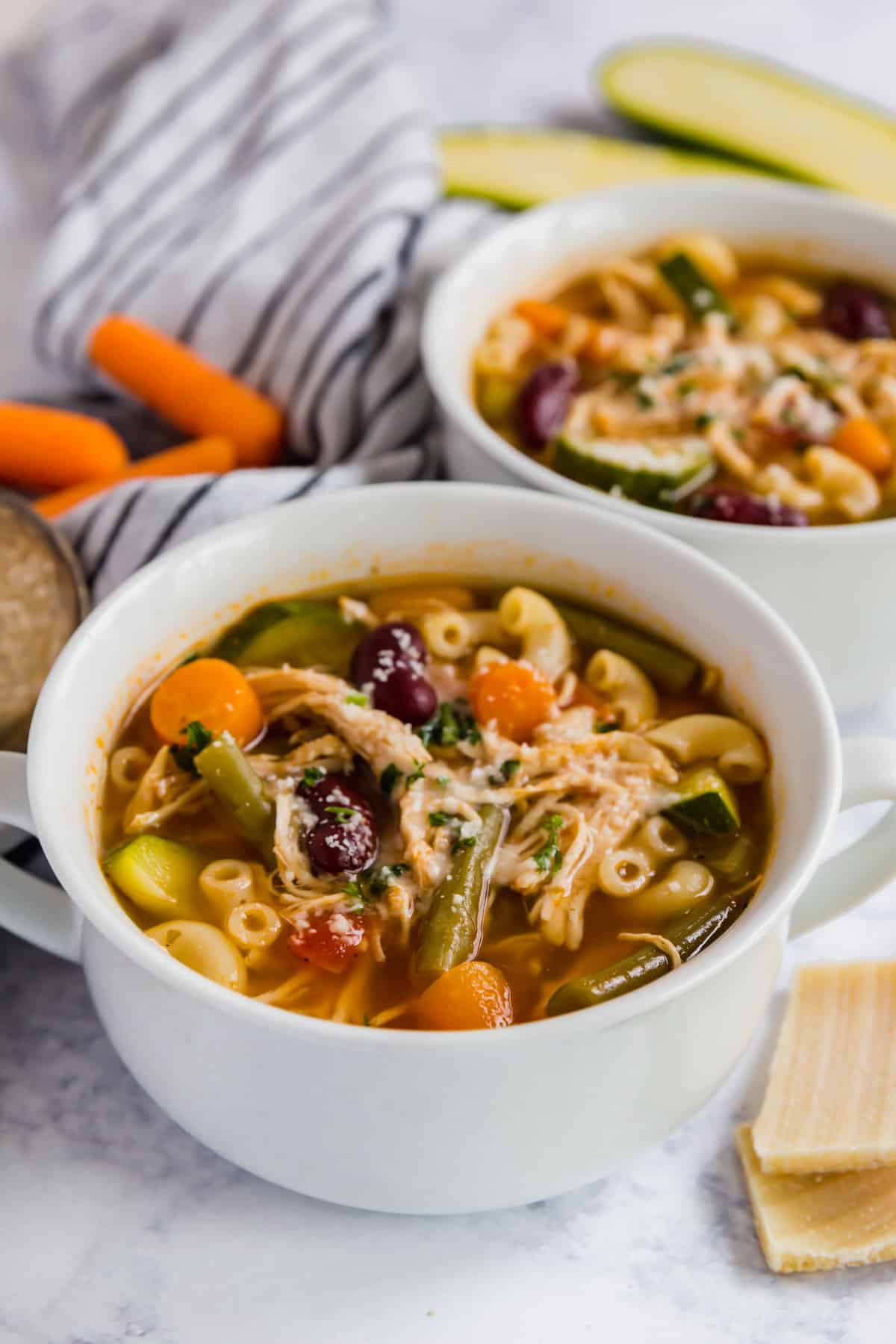 A bowl of minestrone with a second bowl behind it and veggies and Parmesan rinds around it.