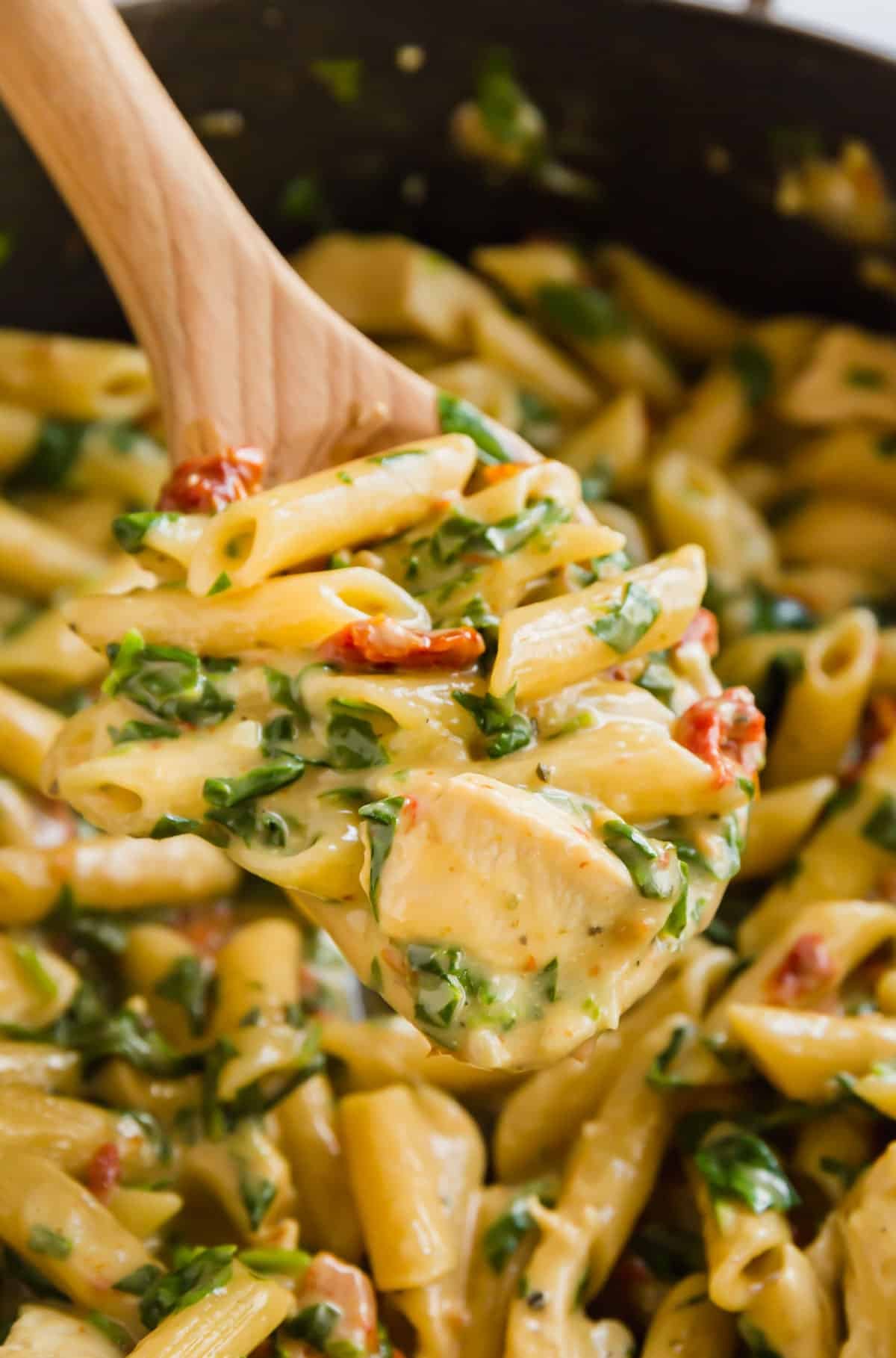 A wooden spoon holding a scoop of creamy chicken Florentine pasta over the full pan of it.