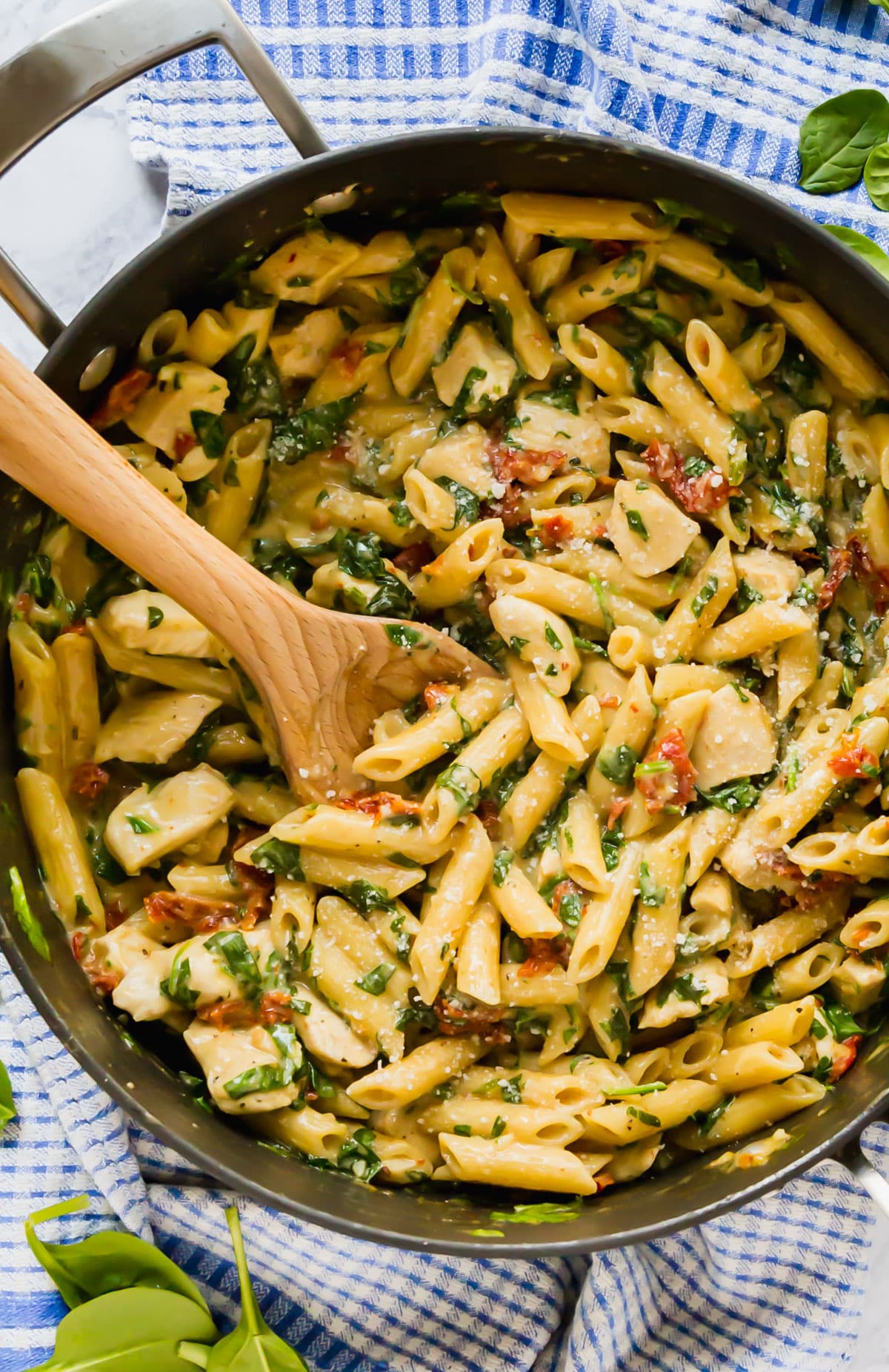 An overhead image zooming in on a pan of chicken and pasta Florentine in a pan with a wooden spoon in it.