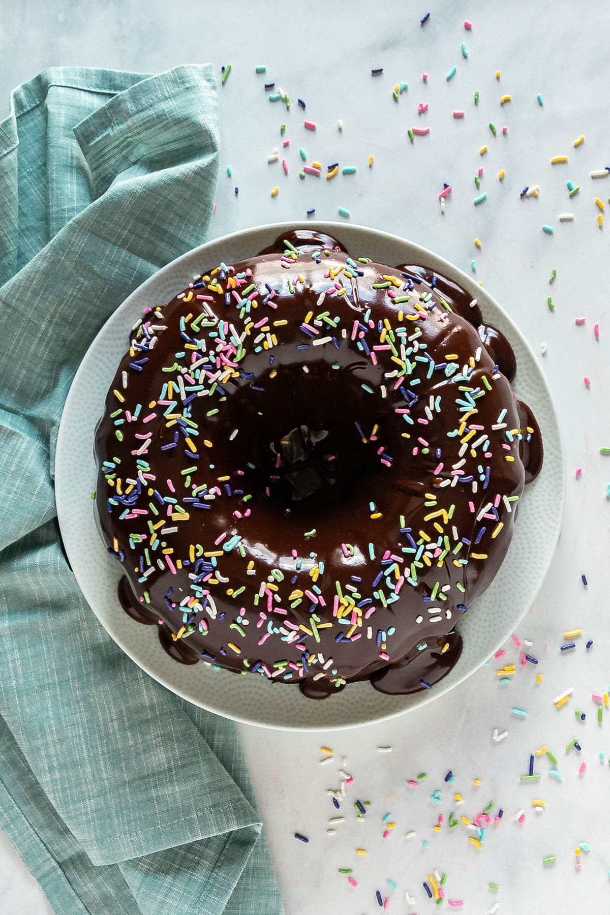 A overhead image of a bundt cake with ganache and sprinkles on top with a teal napkin and other sprinkles on the side.