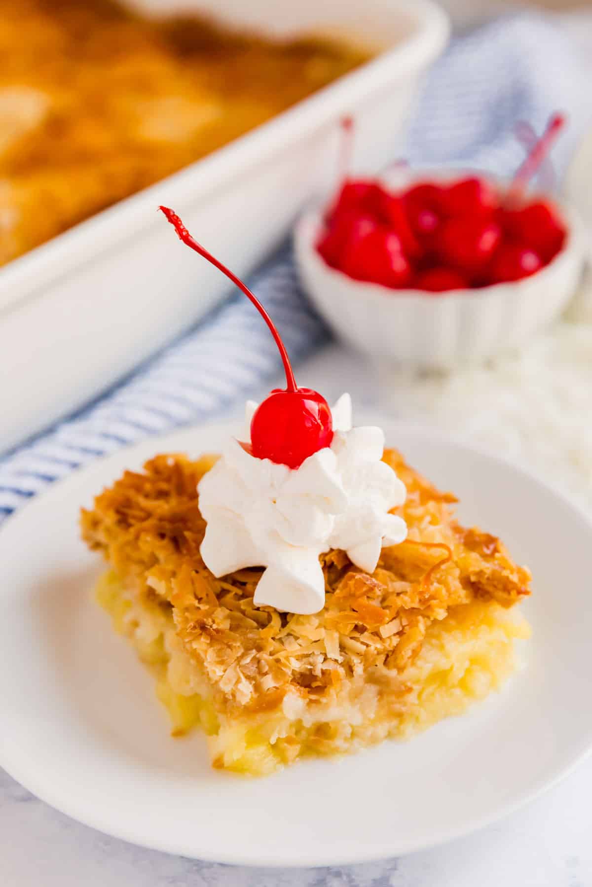 A side angle shot of a pice of dump cake with whipped cream and a cherry on top with a bowl of maraschino cherries and the whole dump cake int he background.