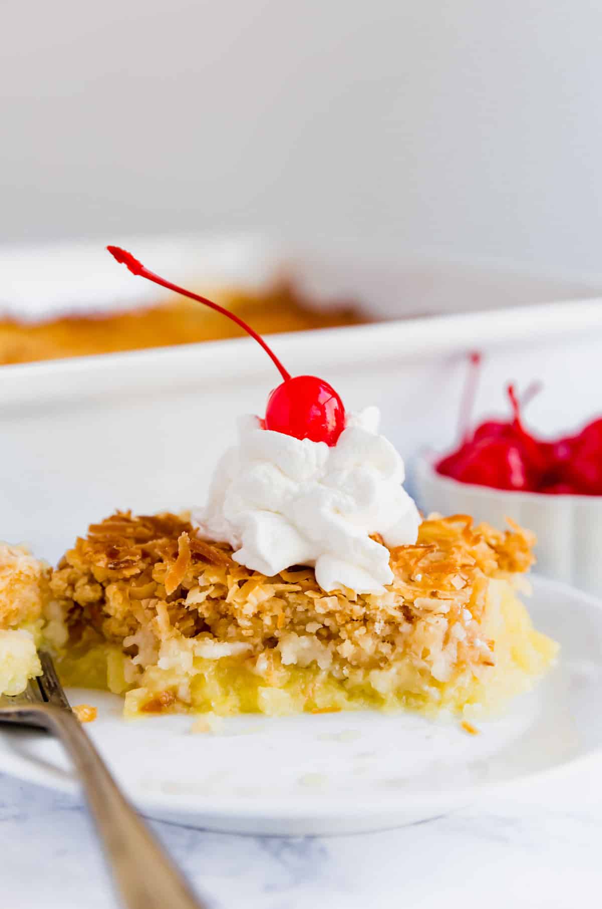 A close up image of Piña Colada dump cake on a plate with whipped cram and a cherry on top on a white plate with a fork.