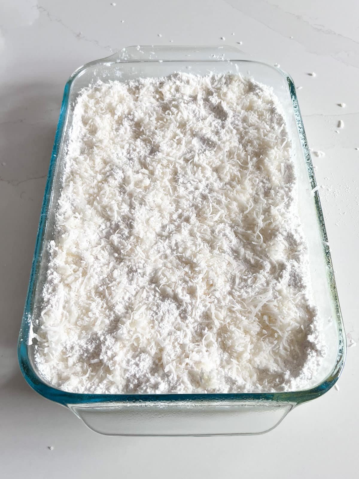 A layer of coconut sprinkled on top of the cake mix layer in a glass baking pan.