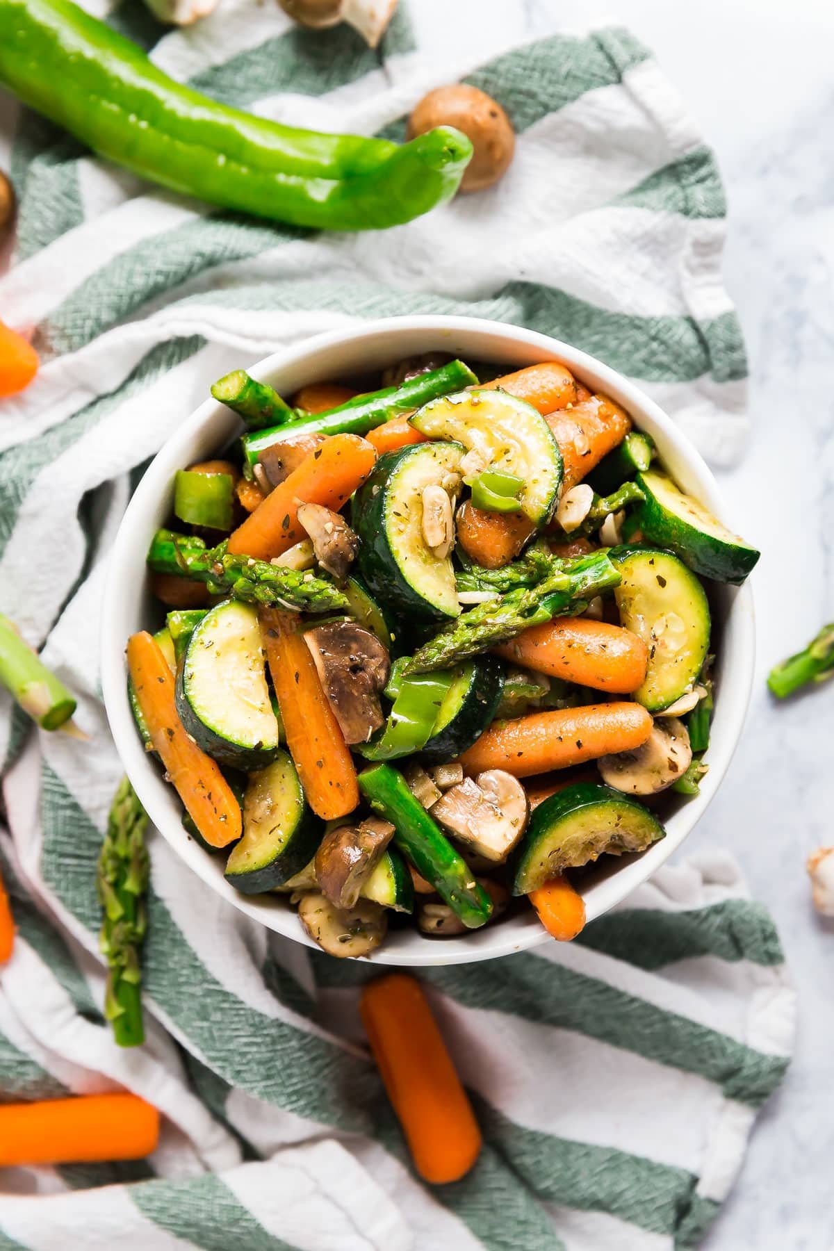 An overhead image of a bowl of sautéed veggies on top of a green and white striped towel with raw veggies around it.
