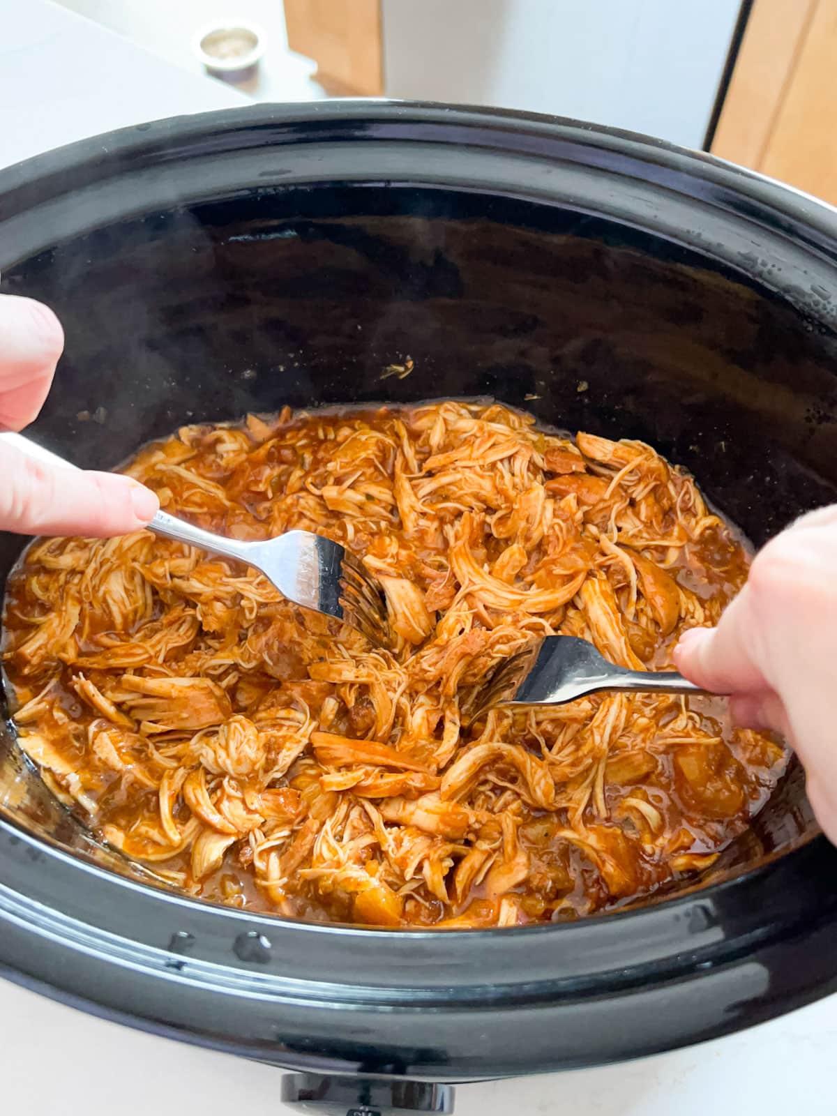 Hands using two forks to shred the cooked chicken in the crockpot.