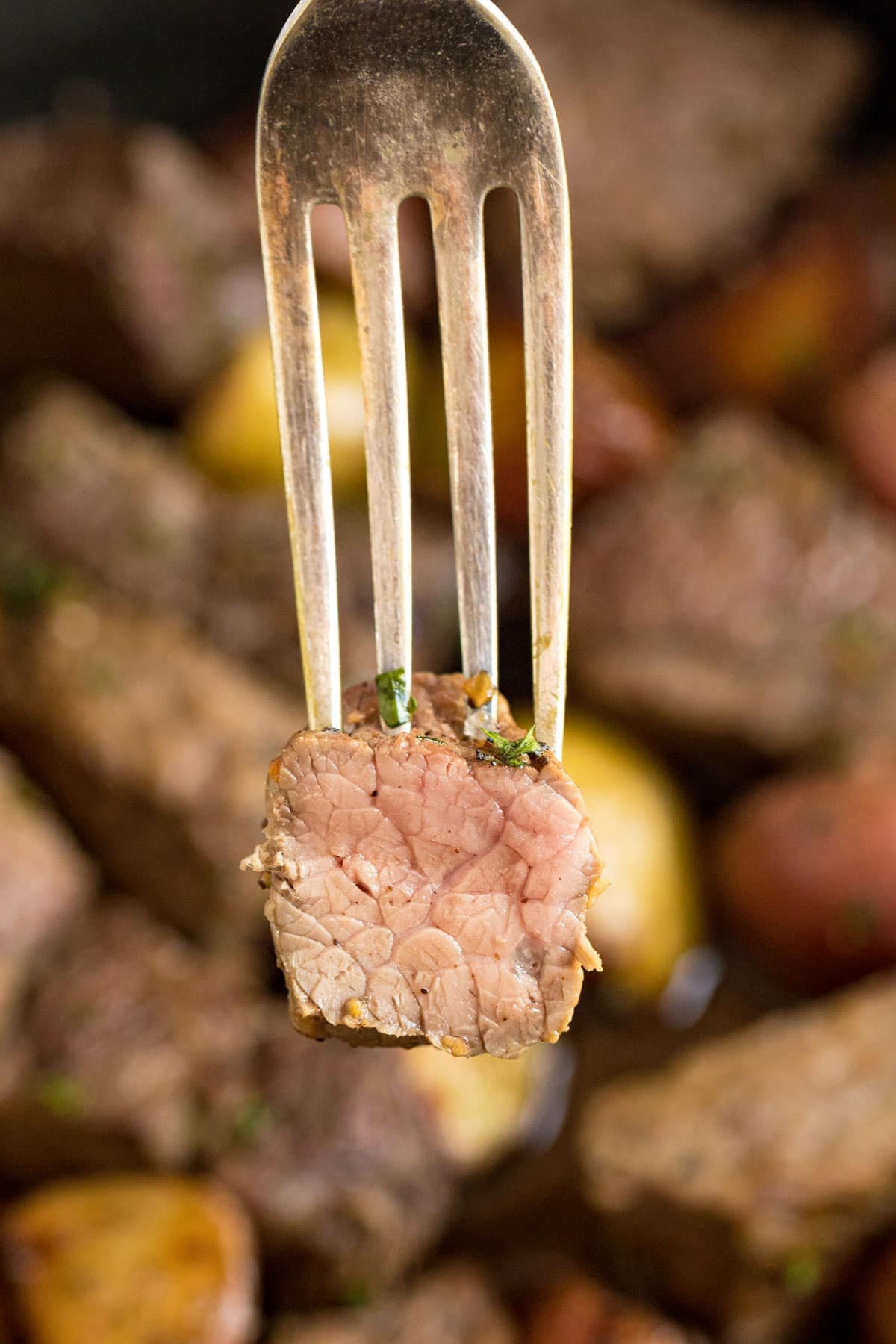A close up image of a fork holding up a cooked piece of steak that's cut revealing the medium inside of the steak. 