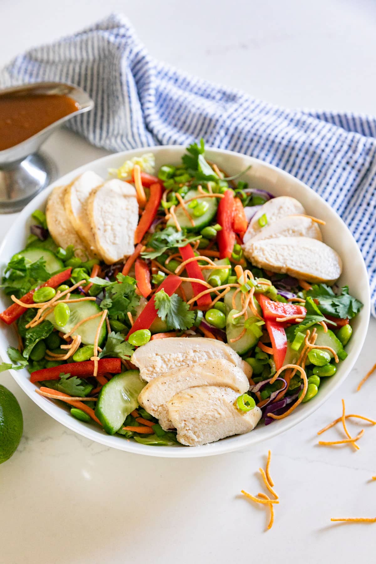 A salad with chicken and rainbow veggies on it in a white bowl with a blue napkin and dressing in the background.