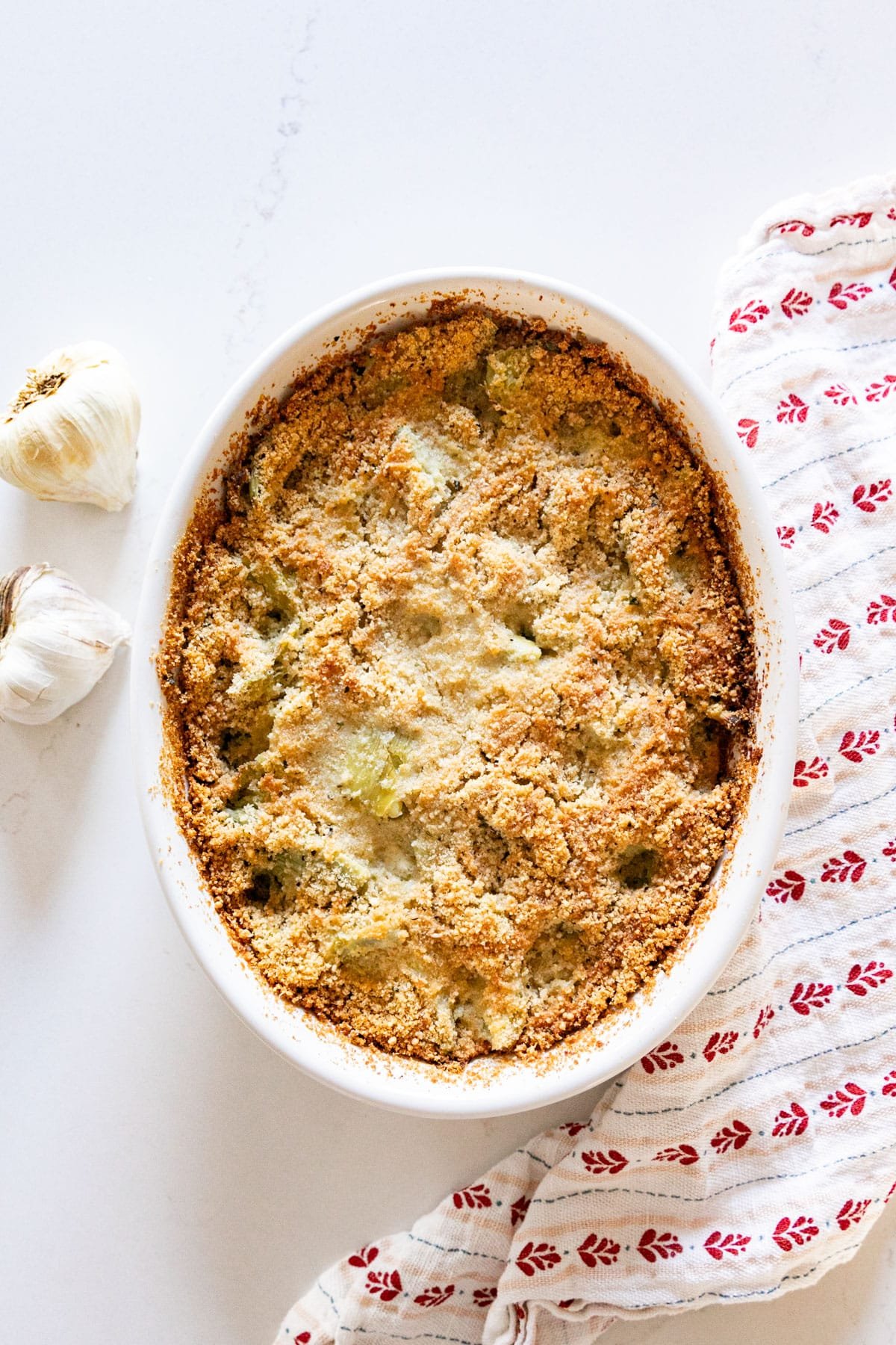 An overhead image of this artichoke bake recipe in a white oval baking dish with a white and red napkin and some garlic next to it.