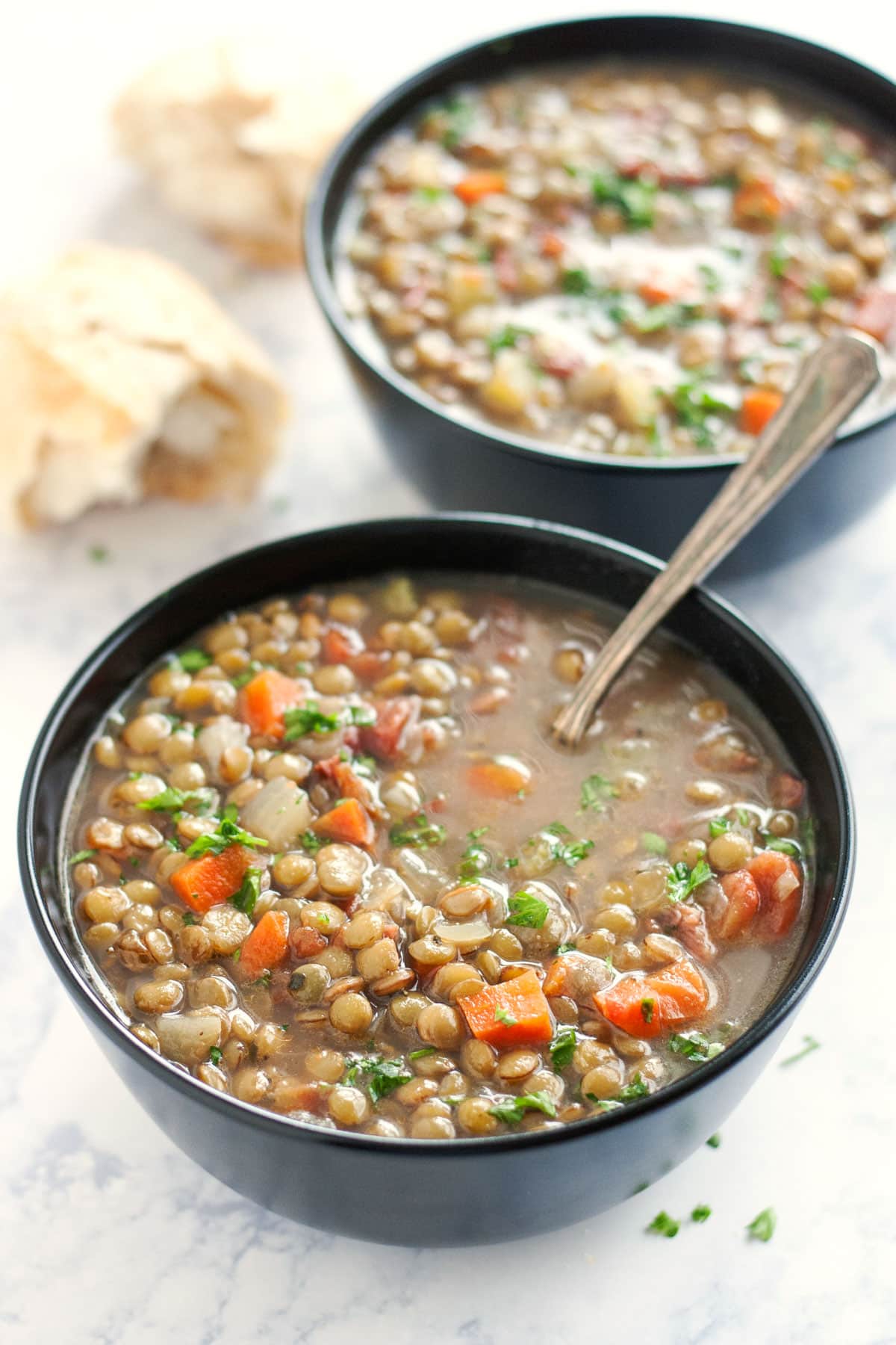 Two black bowls of lentil soup with bread next to it and a spoon in the front bowl.