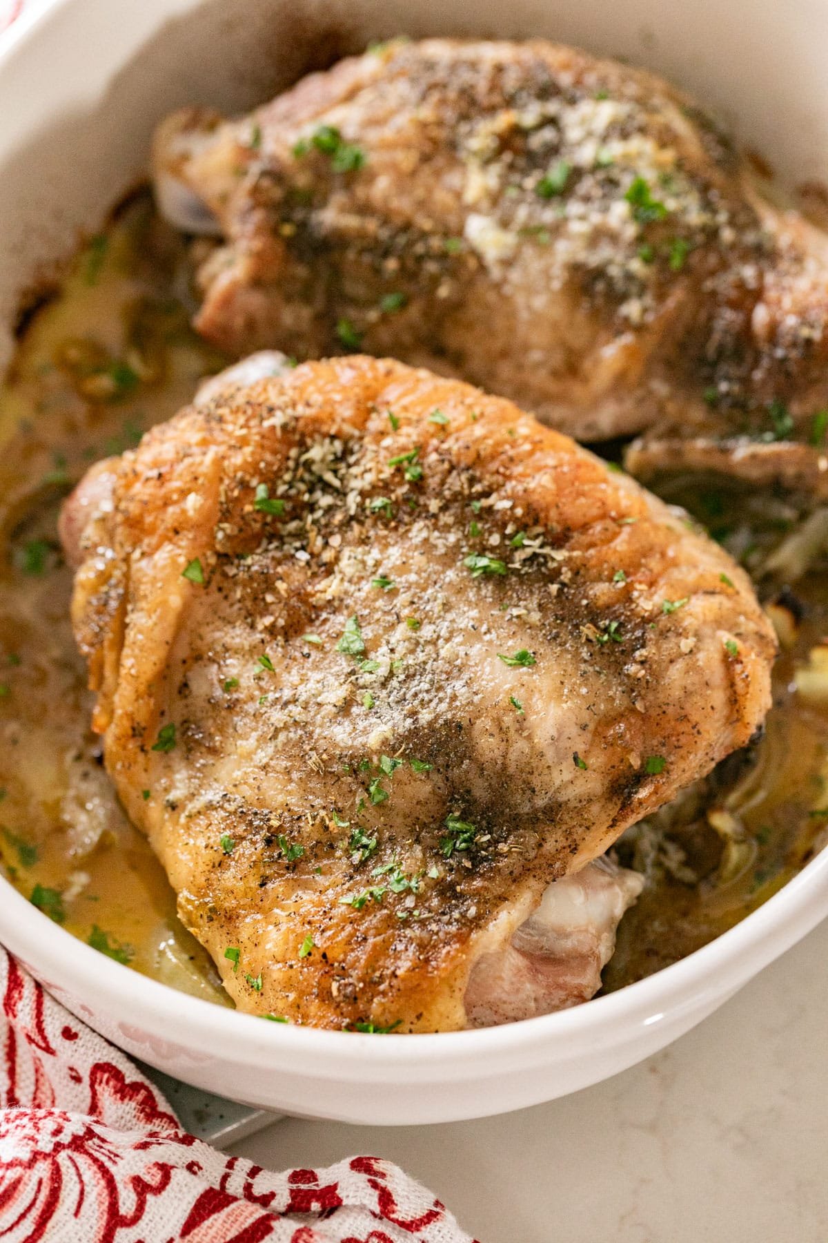 A close up image of golden brown roast turkey thighs in a white pan focusing on one of the turkey thighs with a red and white napkin off to the side.