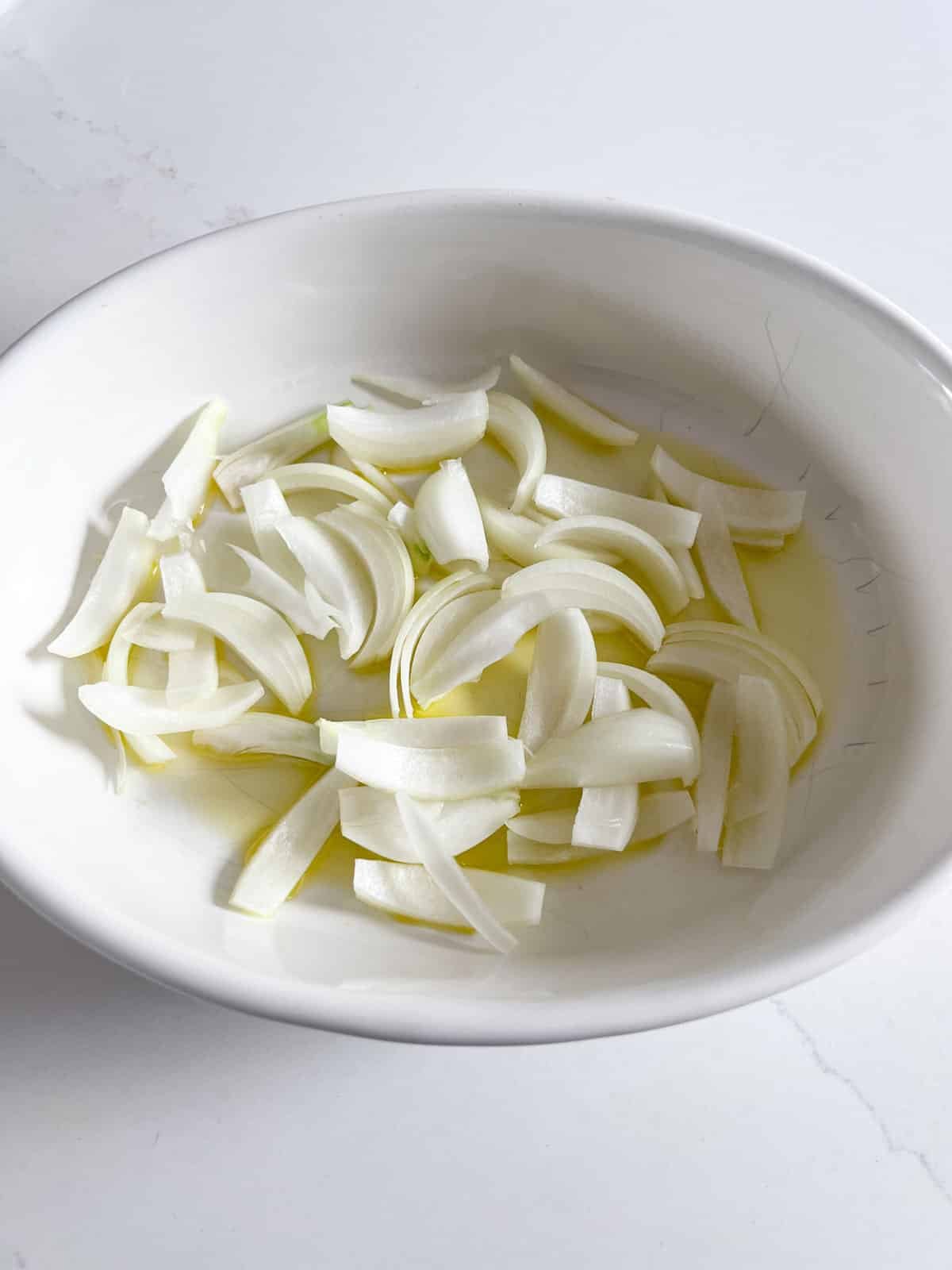 Sliced onions and oil in a white oval baking pan.