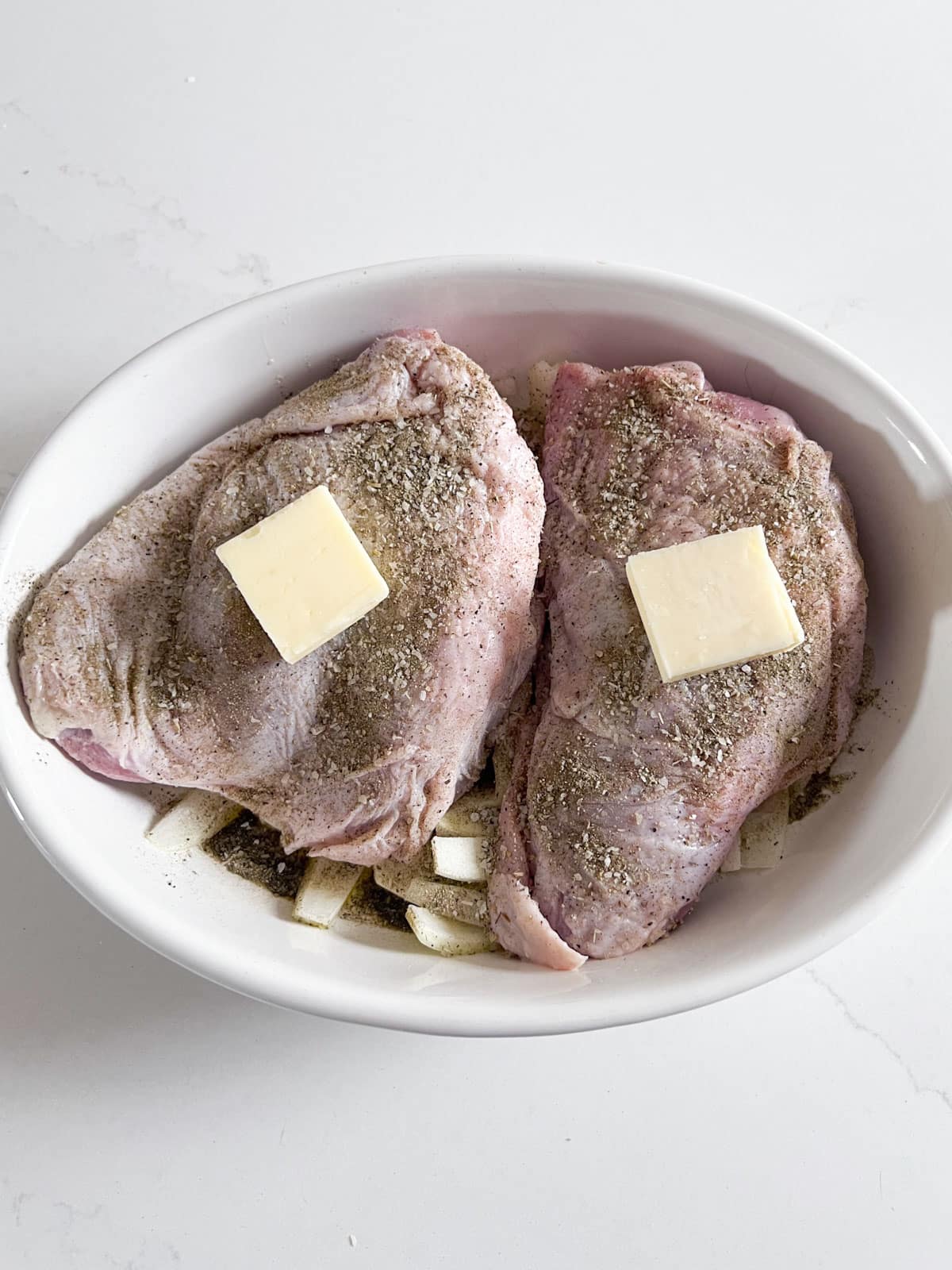 Pats of butter added to the top of the turkey thighs in a white oval baking pan.