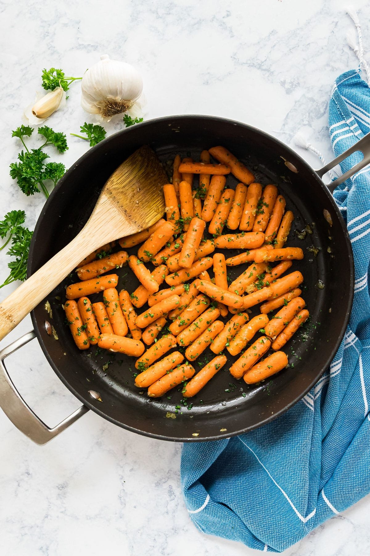 An overhead image of a pan of carrots with a wooden spoon in them and a blue towel, parsley and garlic on the side around it.