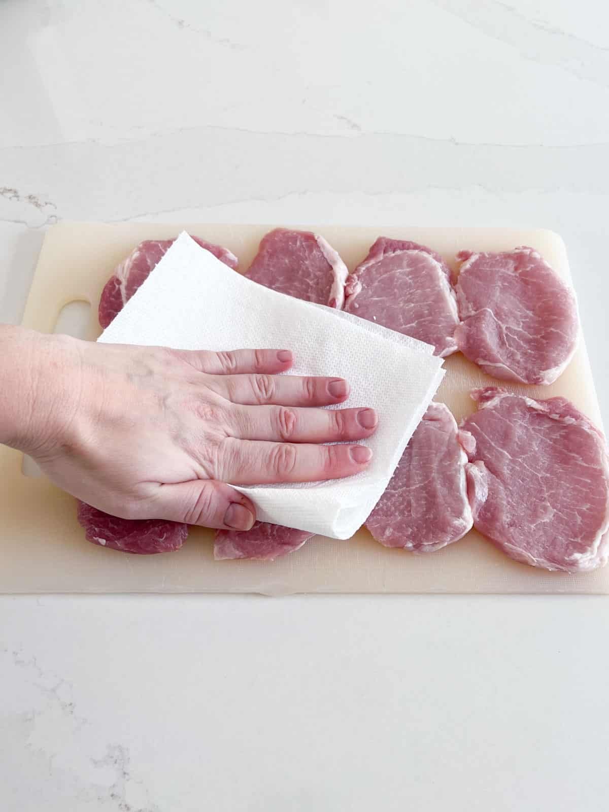 A hand patting dry thin pork chops with a paper towel on a white cutting board.