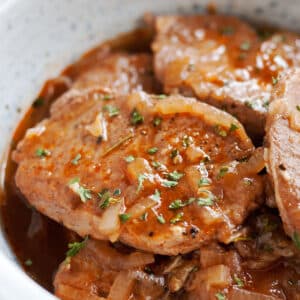 A close up image of a red wine braised pork chop with onions on it in a gray bowl of other pork chops and sauce.
