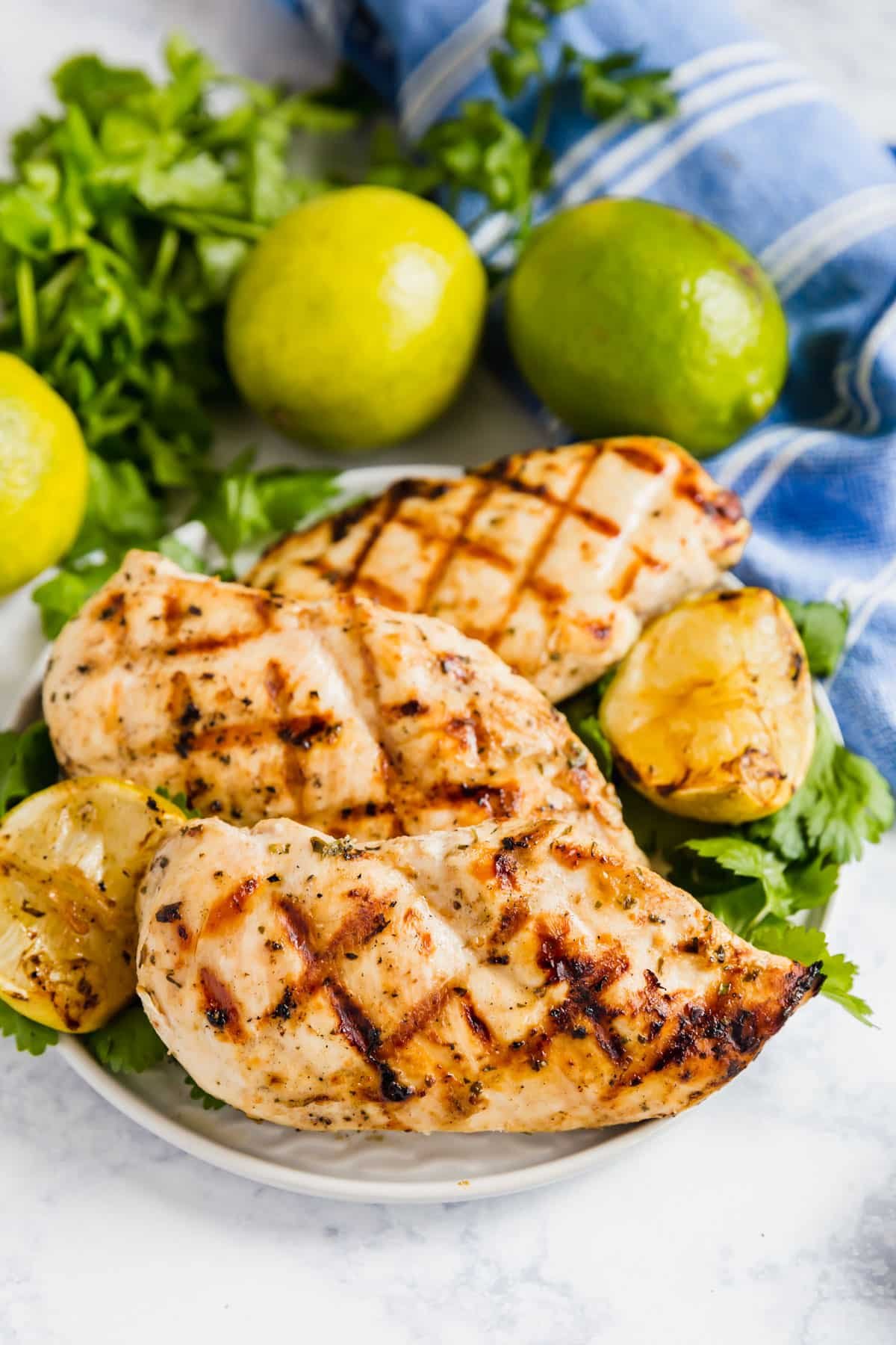 coconut lime grilled chicken breasts on a plate with cilantro and limes behind it and a blue striped napkin.