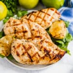 Coconut lime grilled chicken breasts on a plate with cilantro and limes behind it and a blue striped napkin.
