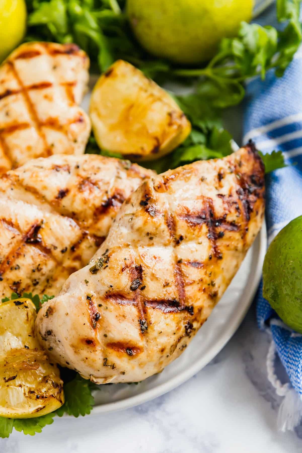 Focusing on a grilled chicken breast on the side of a plate of chicken with cilantro and limes in the background.