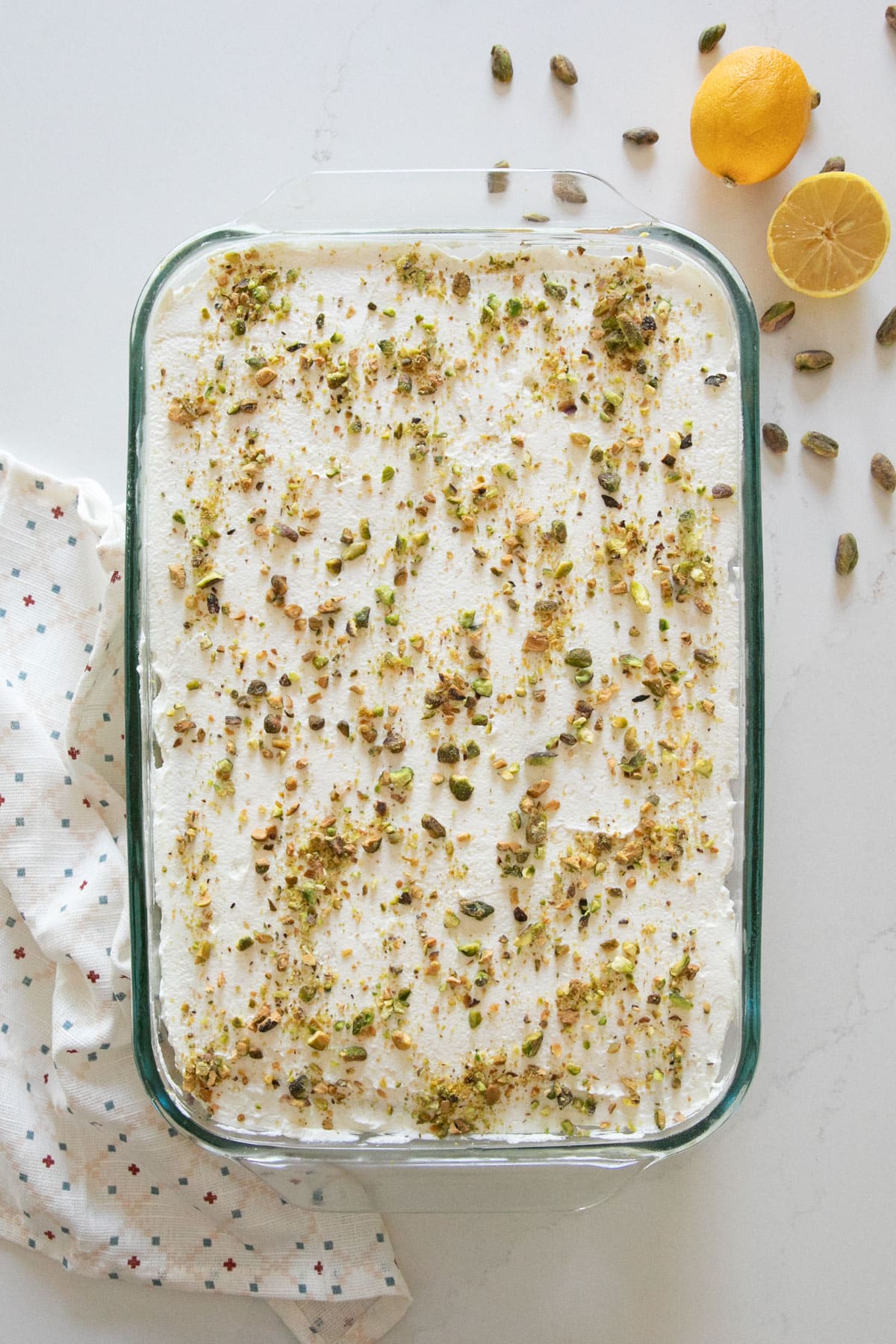 An overhead image ofa pistachio cake with whipped cream and sprinkled with pistachios in a rectangular glass pan with a white towel, pistachio nuts and lemons on the sides of it.