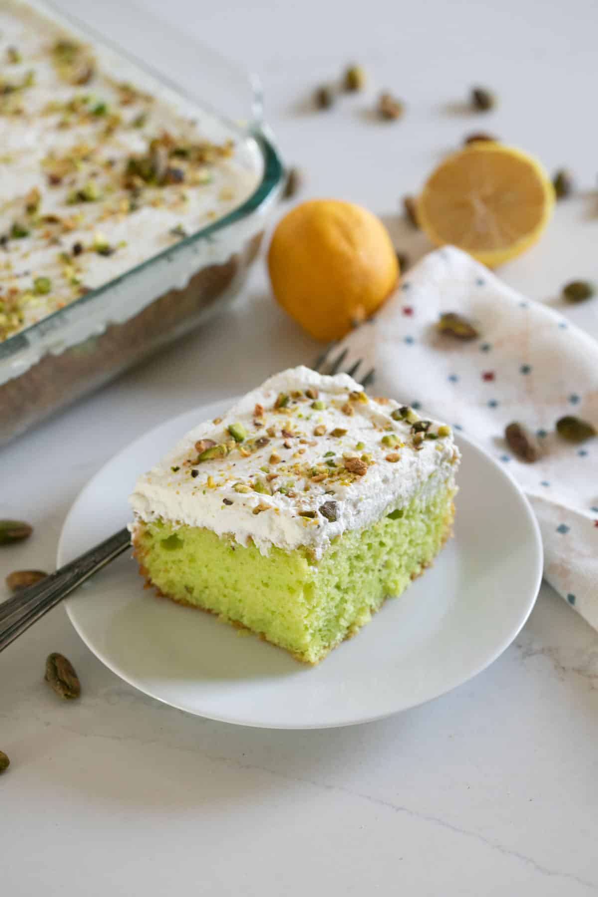 A slice of lemon pistachio cake on a white plate  with the full cake, lemons and pistachios in the background on a white napkin.