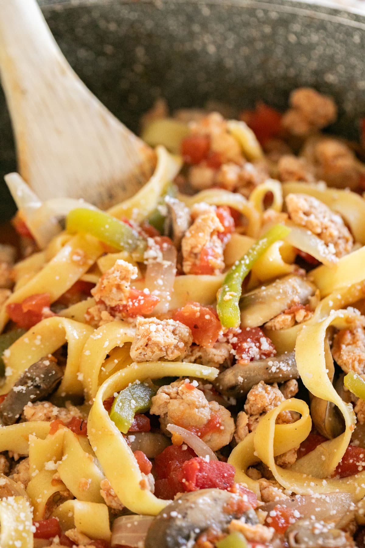 A close up of pasta with tomatoes, peppers and onions ad sausage in a pan with a wooden spoon in it.