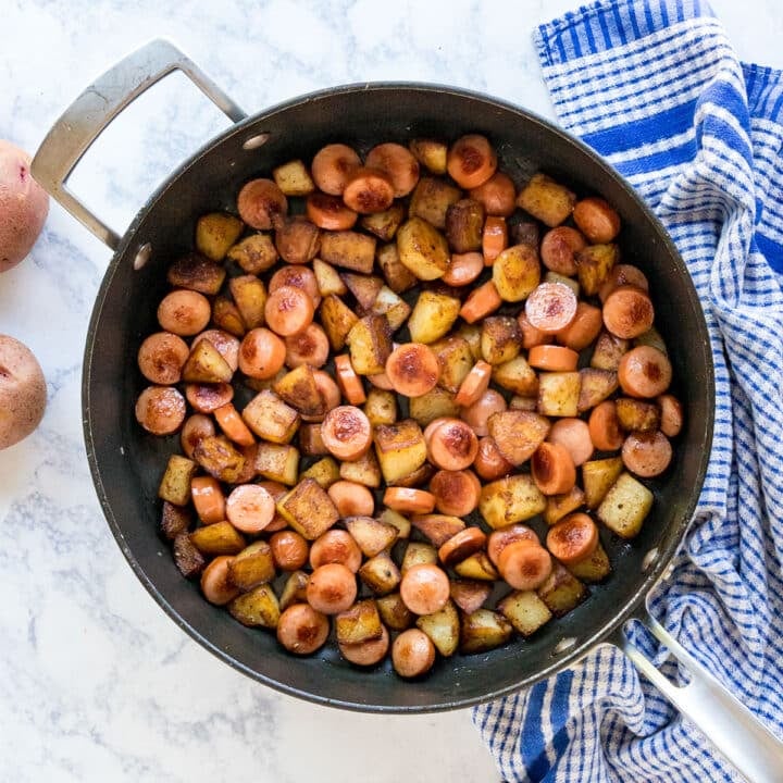 An overhead image of a pan of hot dogs and potatoes with a blue towel and potatoes next to it.