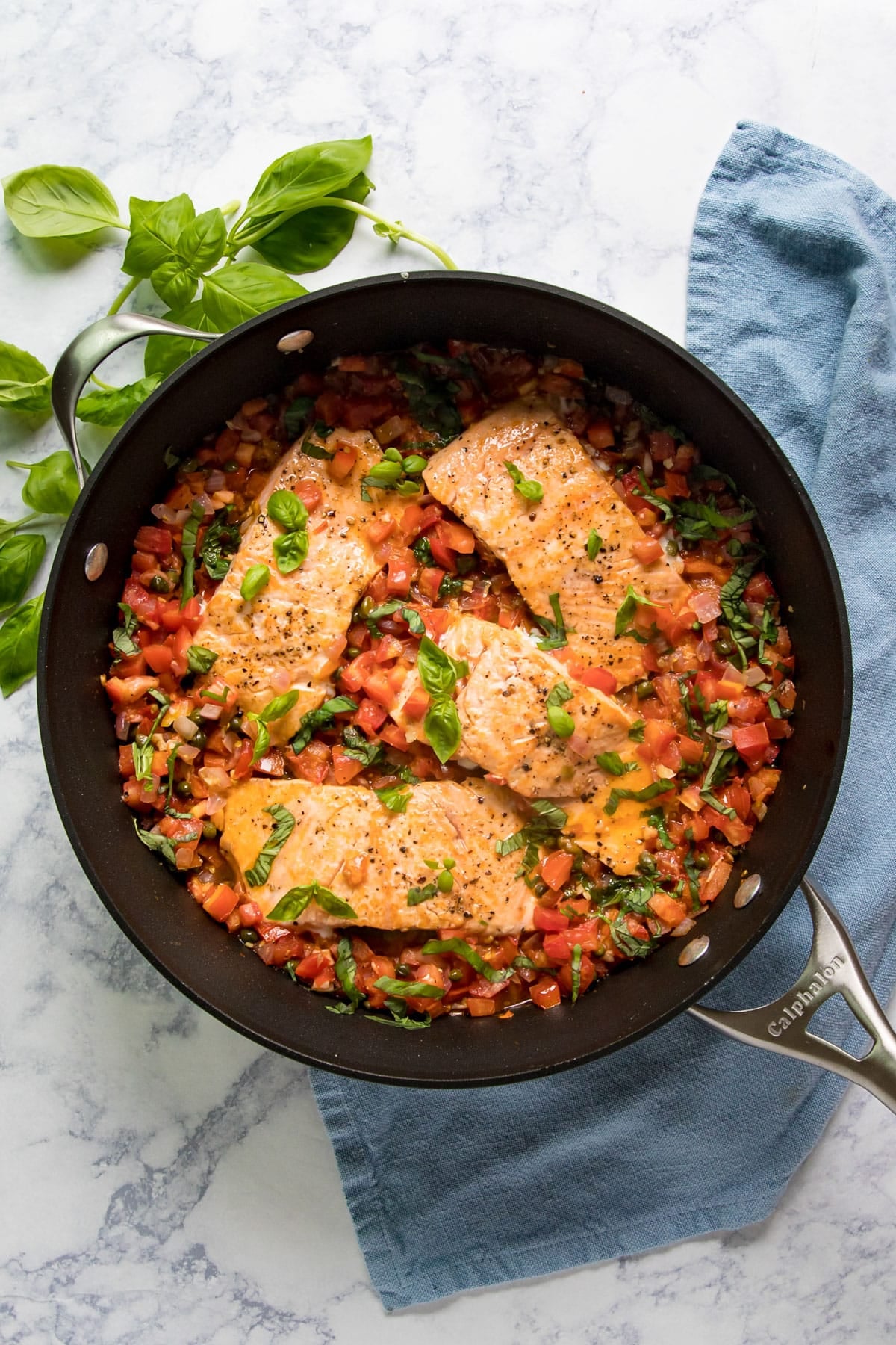 An overhead image of palmon filets in a pan with capers and tomatoes and a blue napkin and fresh basil around it.