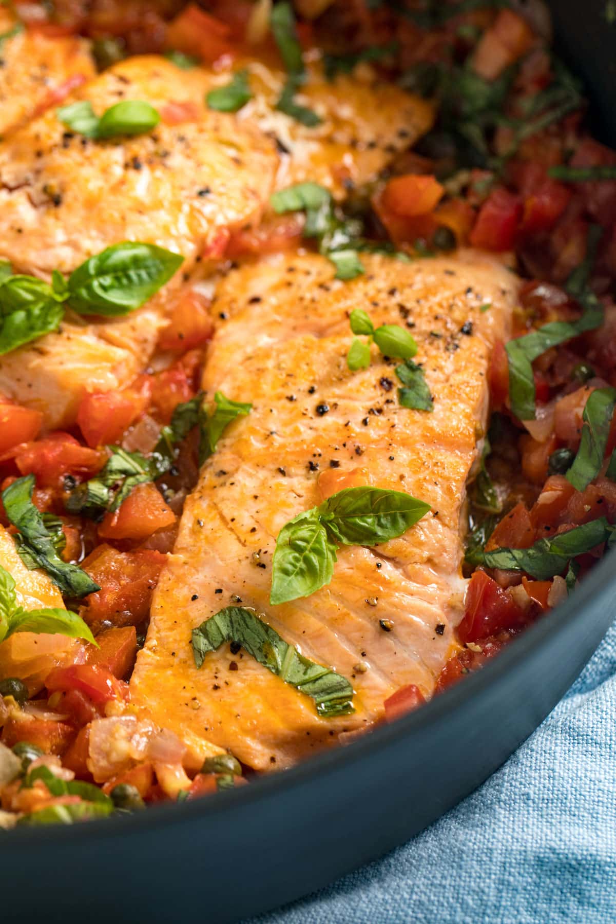 A close up image of a salmon filet nestled in a pan full of tomatoes and capers.