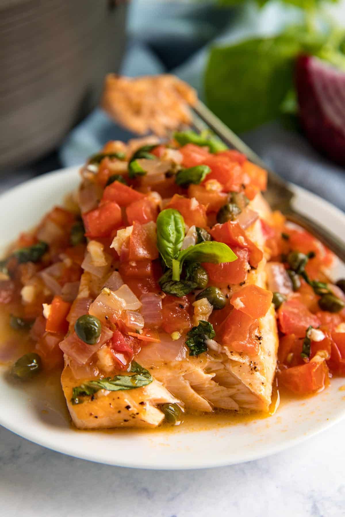 A close up image of salmon in tomato and caper sauce with a bite taken from it revealing the perfectly cooked inside of the salmon filet.